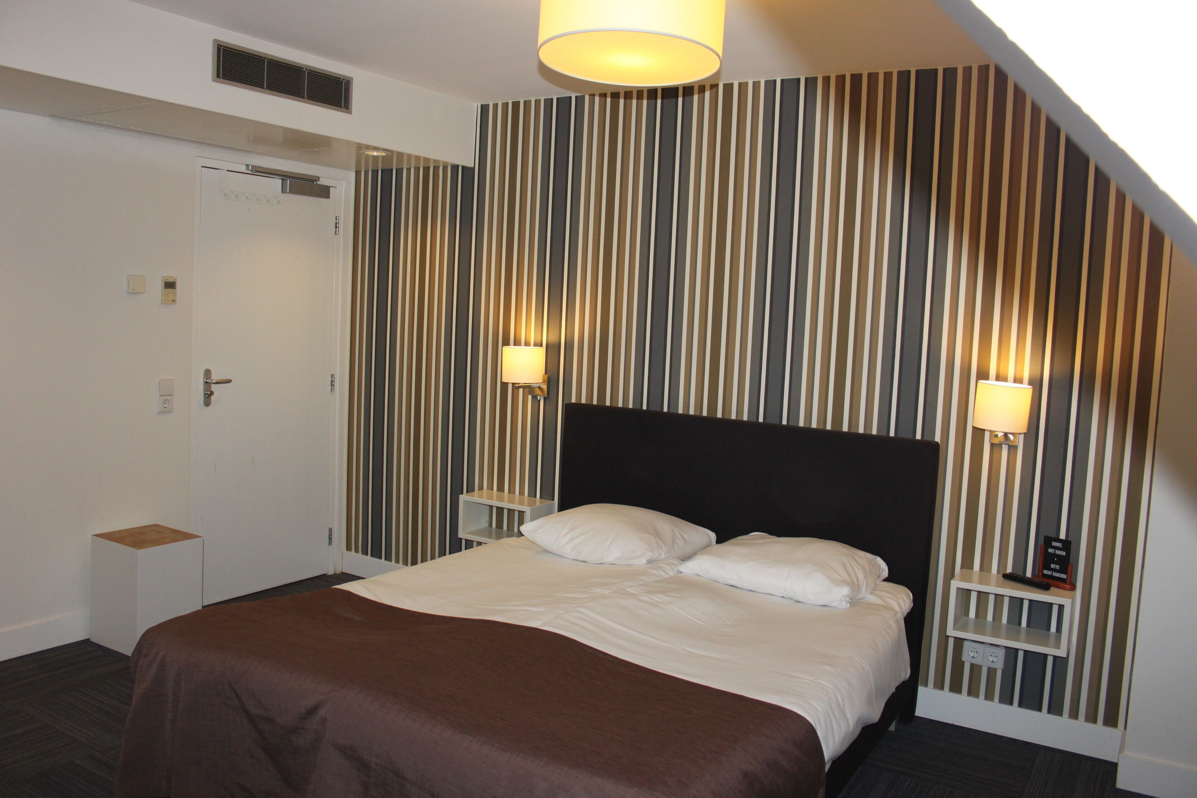 Une chambre d’hôtel avec un lit, deux tables de chevet, une petite table, une lampe et un mur à rayures.