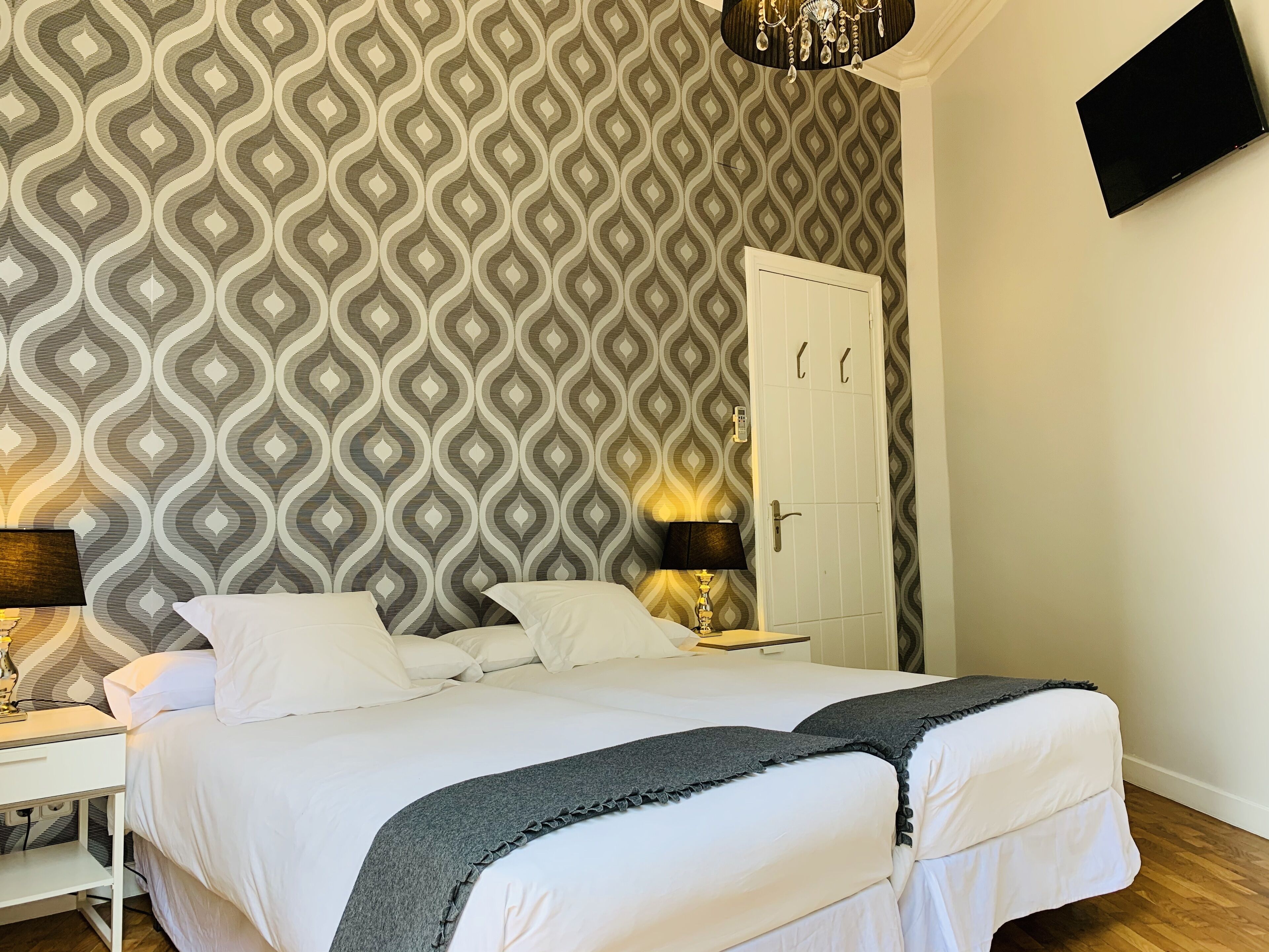 Hotellihuone, jossa on kuviollinen tapetti, valkoisilla lakanoiden ja harmaan peiton peitetty sänky, yöpöytä lampun kanssa ja vaatekaappi.