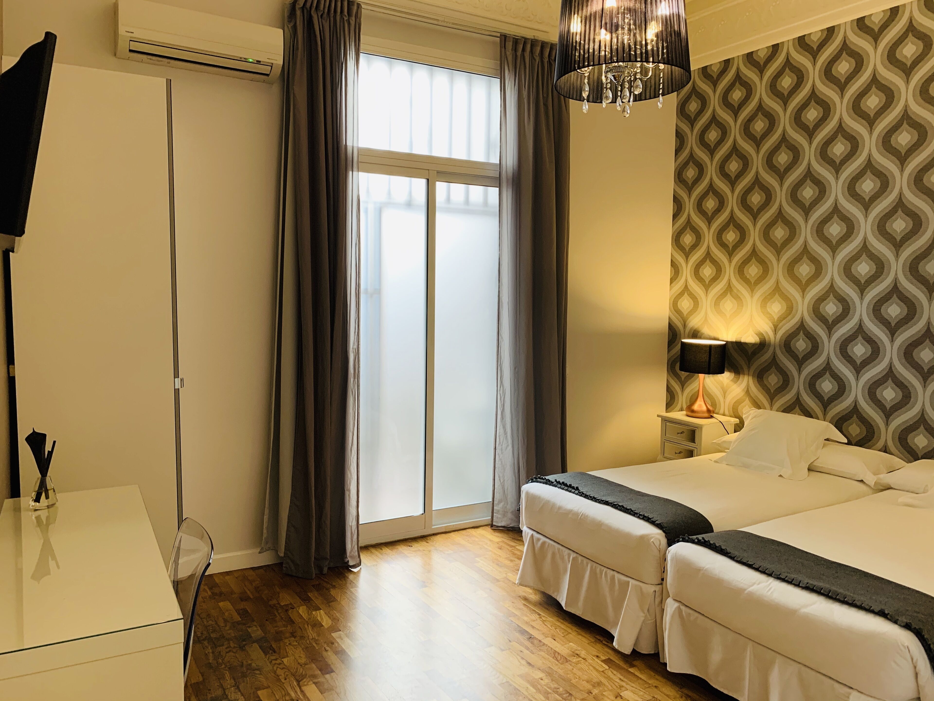 Habitación de hotel con cama, escritorio, silla, lámpara, pared con diseño y ventana con cortinas.