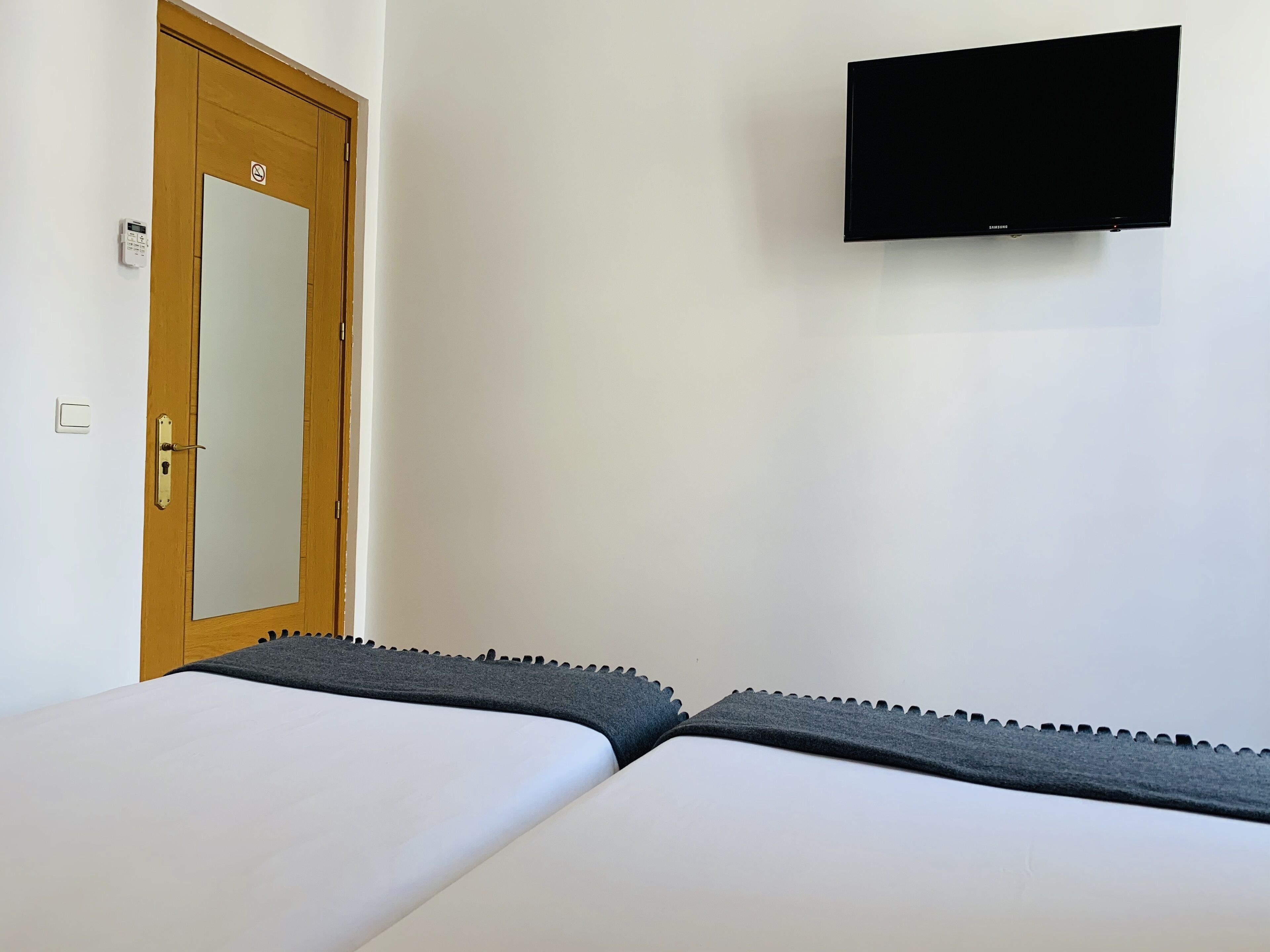 Ein Zimmer mit zwei Betten, einer Holztür mit Verbotsschild und einem an der Wand montierten Fernseher sowie einem Lichtschalter.