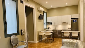 Una cocina moderna con armarios blancos, un refrigerador de acero inoxidable y un microondas. Hay un comedor con un sofá y sillas blancos.