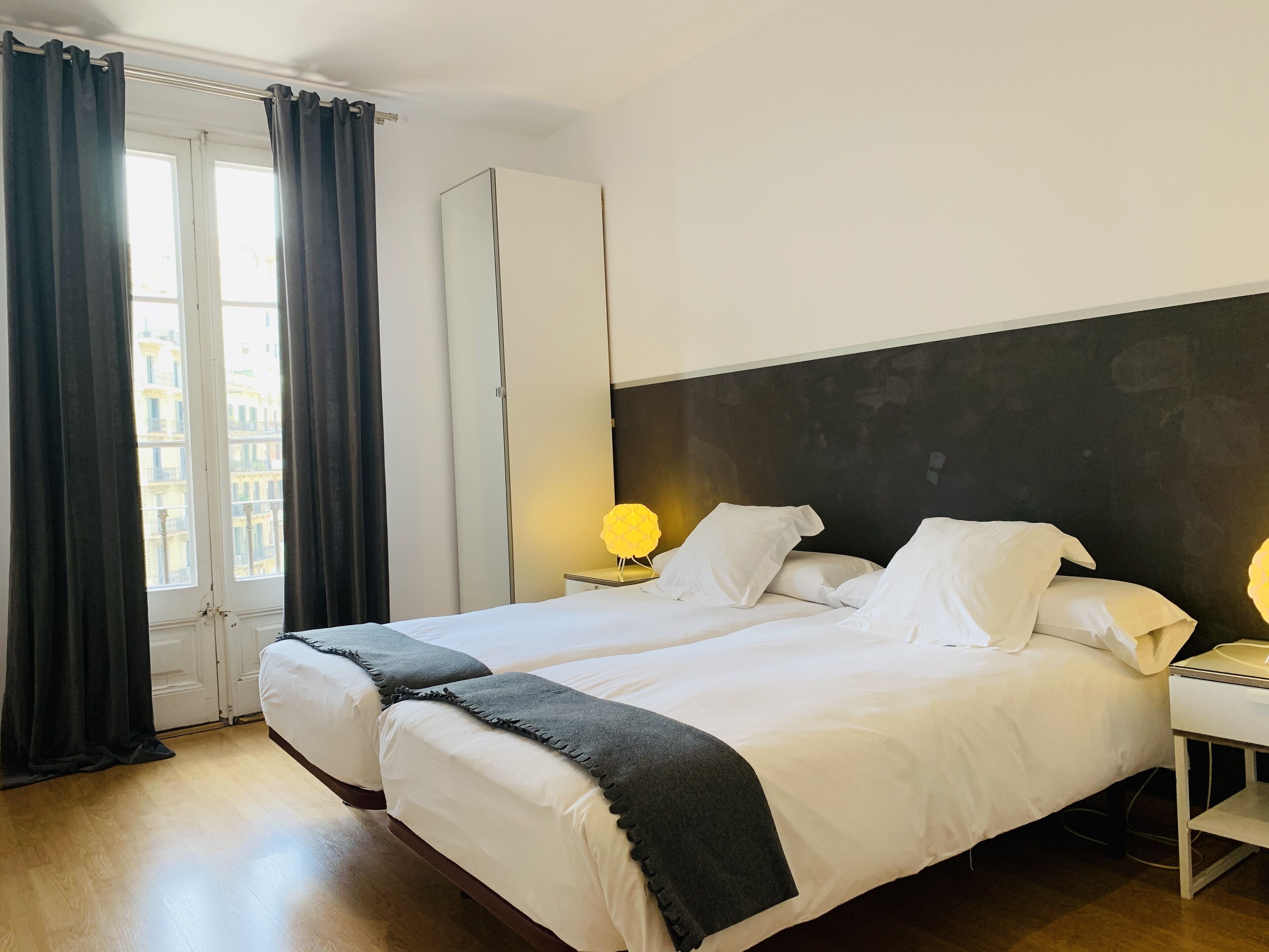 Habitación de hotel con dos camas, un armario, una mesita de noche con lámpara y vistas a edificios desde la ventana.