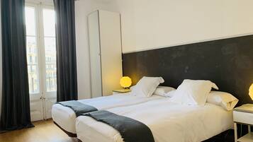Habitación de hotel con dos camas, un armario, una mesita de noche con lámpara y vistas a edificios desde la ventana.