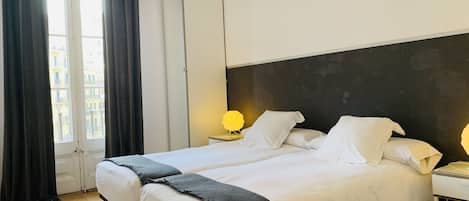 Habitación de hotel con dos camas, un armario, una mesita de noche con lámpara y vistas a edificios desde la ventana.
