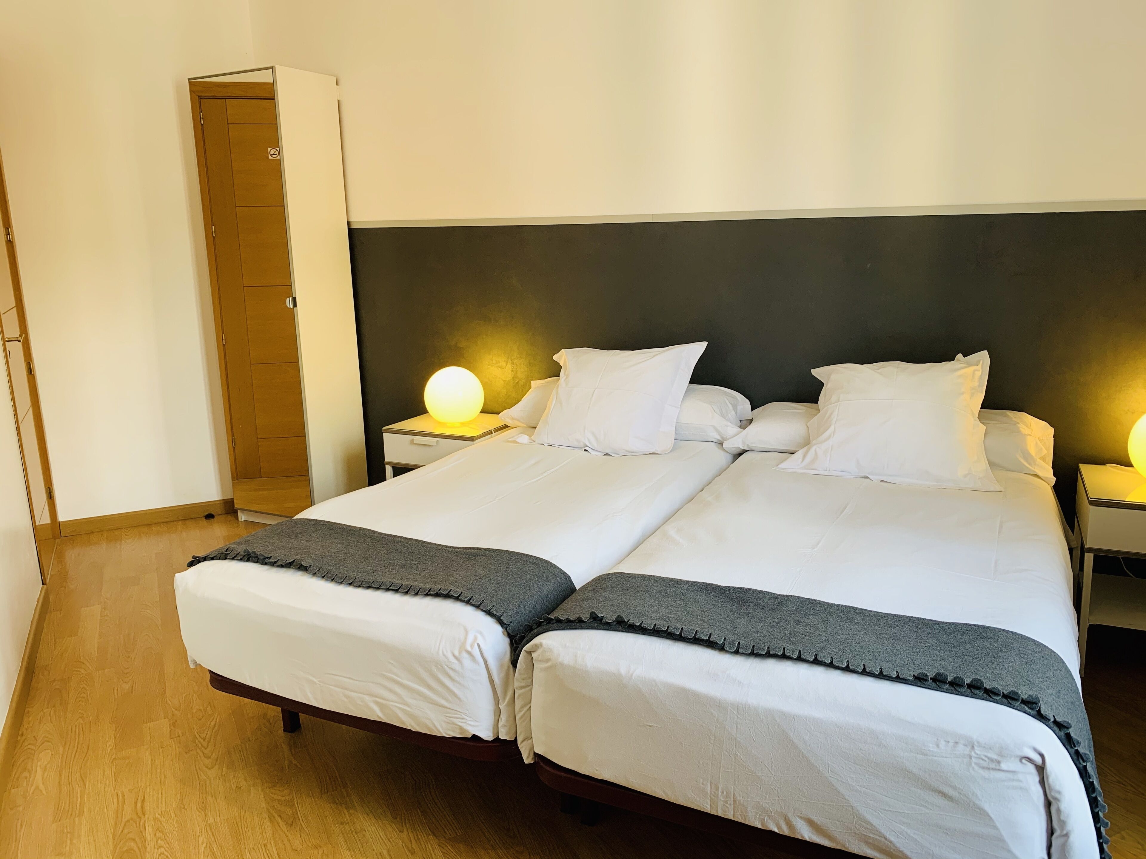 Cama doble con ropa de cama blanca y una manta gris, dos mesitas de noche con lámparas, un armario de madera y una pared lisa.