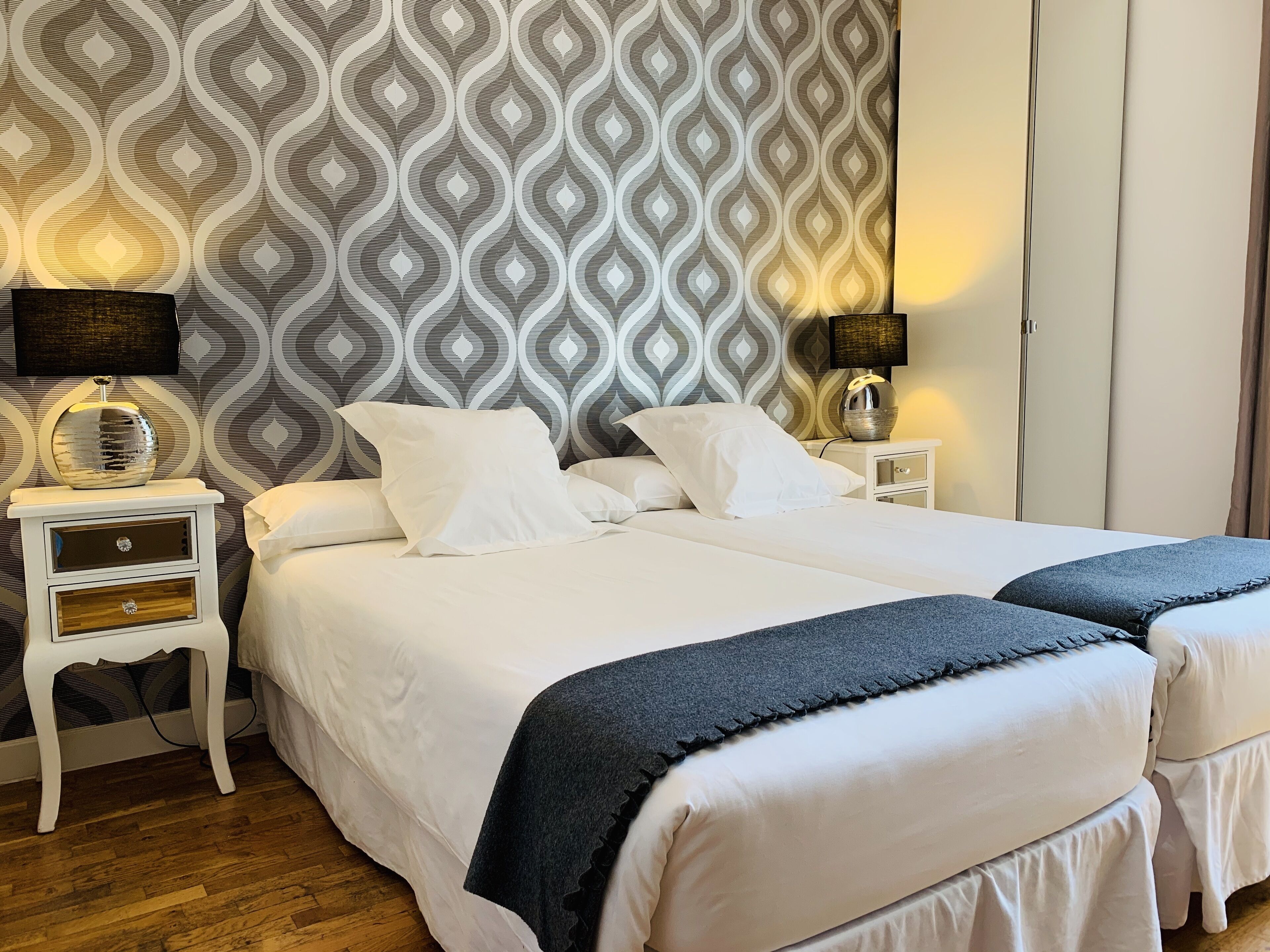 Een keurig opgemaakt tweepersoonsbed met witte lakens en twee kussens, een nachtkastje met een lamp en behang met een patroon.