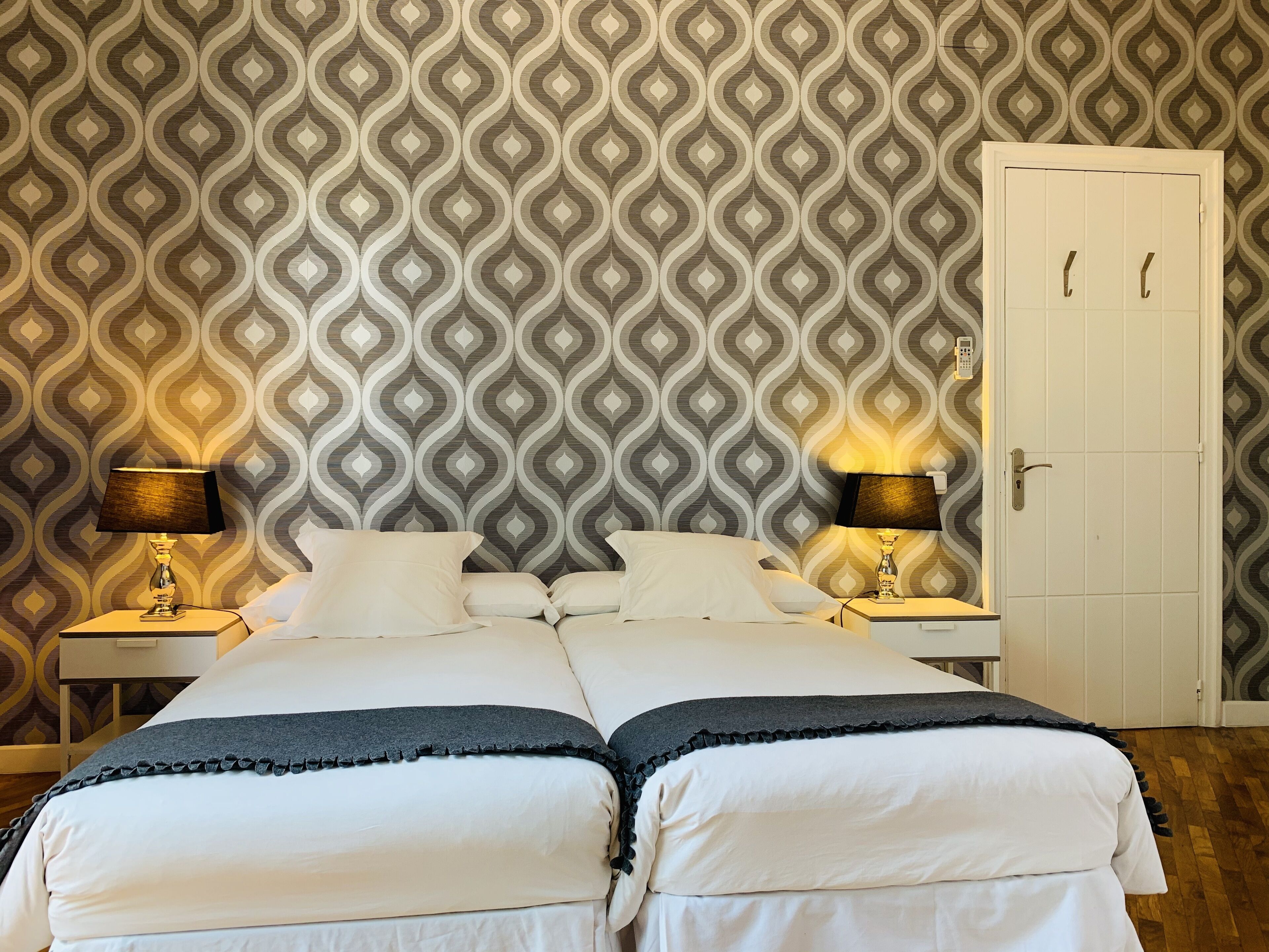 A double bed with two bedside tables, each with a lamp, in a room with patterned wallpaper and a white door.