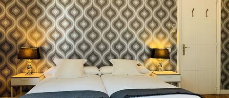 A double bed with two bedside tables, each with a lamp, in a room with patterned wallpaper and a white door.