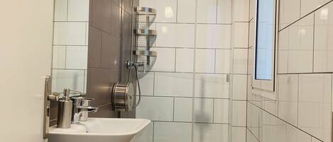 A modern bathroom with a white sink, toilet, and shower. There is a towel rack with folded towels and a small wooden shelf.