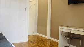 Una habitación con piso de madera, un escritorio blanco con silla, un televisor montado y una puerta que da a otra habitación.