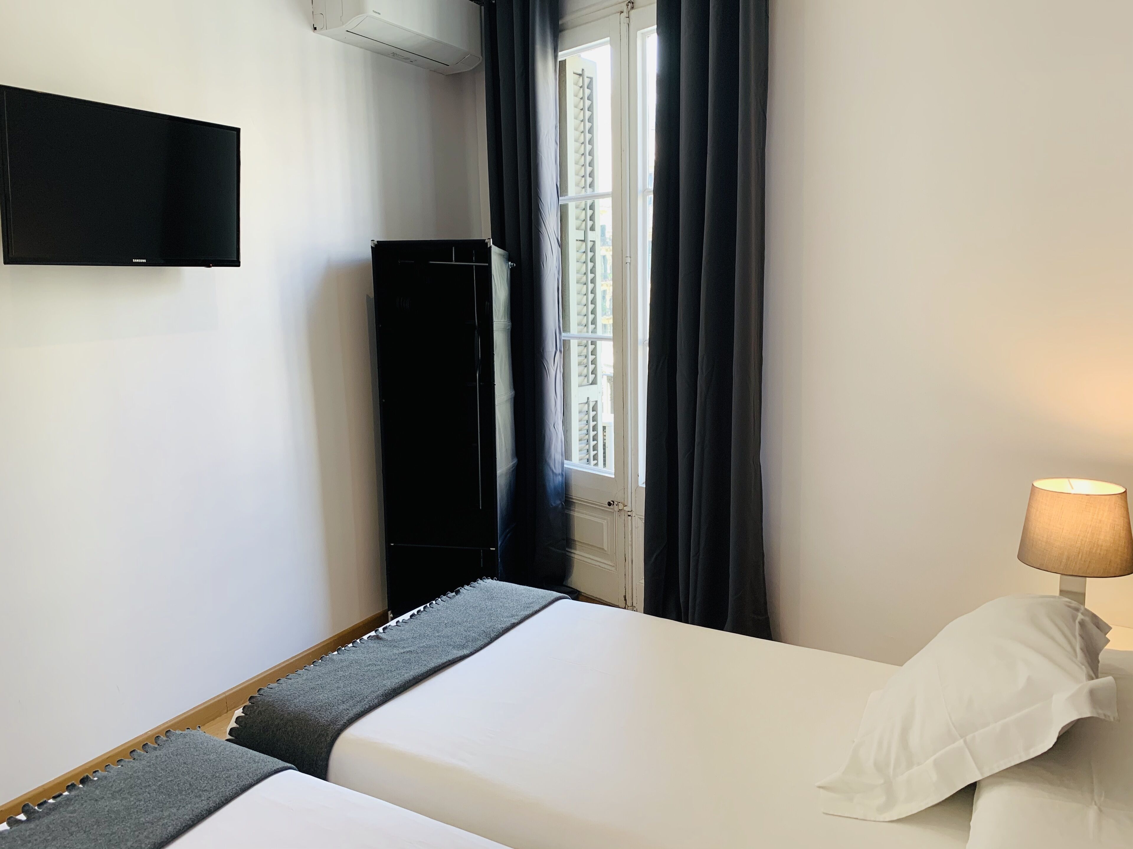 Una camera d'albergo con un letto, una televisione a parete, una finestra con tende e una lampada sul comodino.