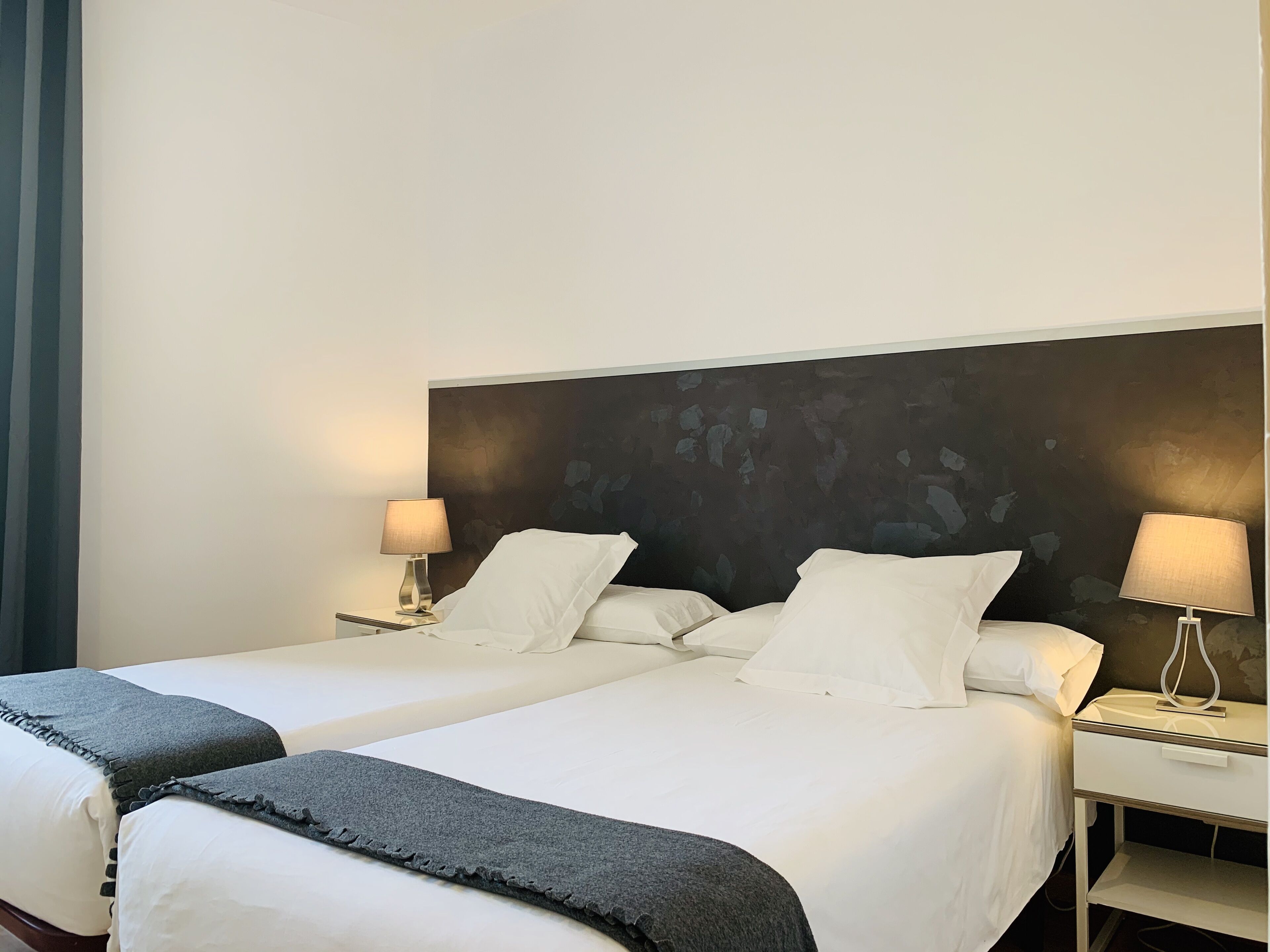 A hotel room with two beds, each with white linens and a dark gray throw blanket. There are two bedside tables with lamps, and a large abstract painting on the wall behind the beds.
