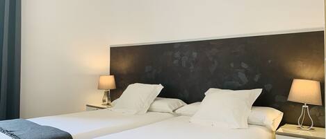 A hotel room with two beds, each with white linens and a dark gray throw blanket. There are two bedside tables with lamps, and a large abstract painting on the wall behind the beds.