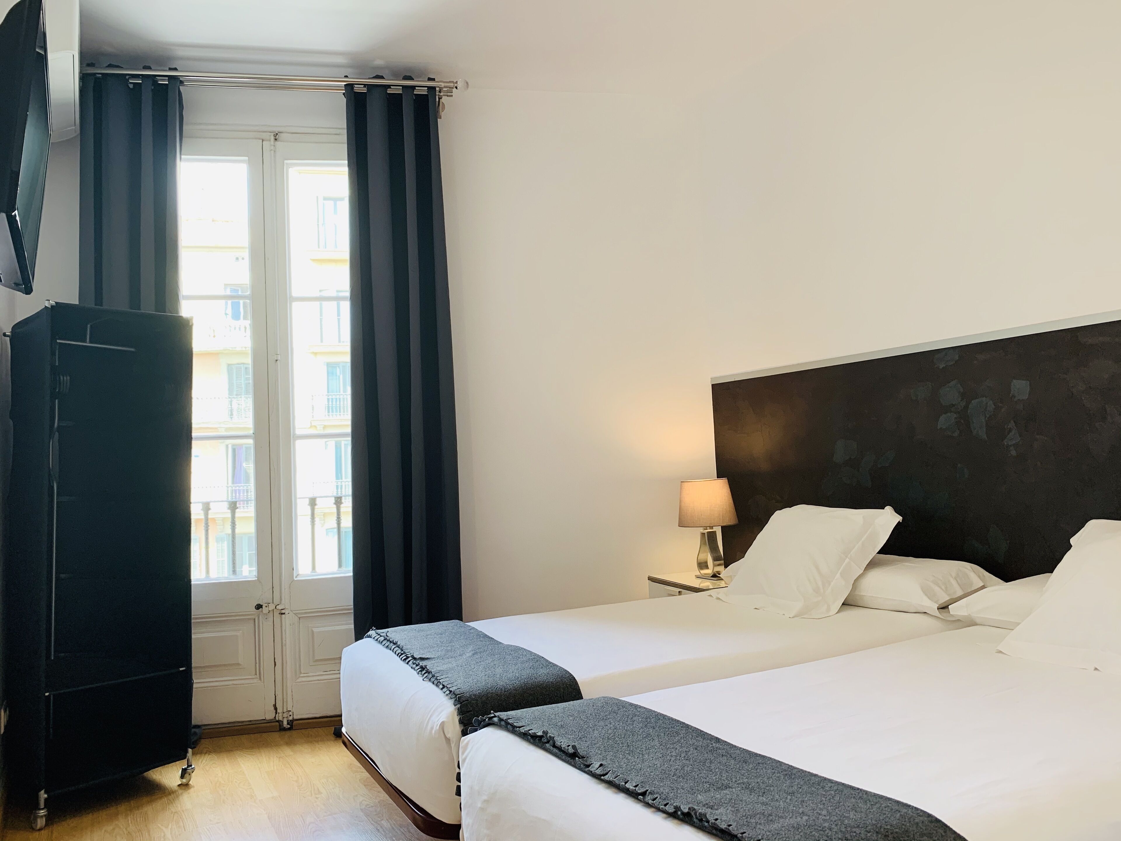 Habitación de hotel con dos camas, un armario, una lámpara y vistas a edificios a través del ventanal.