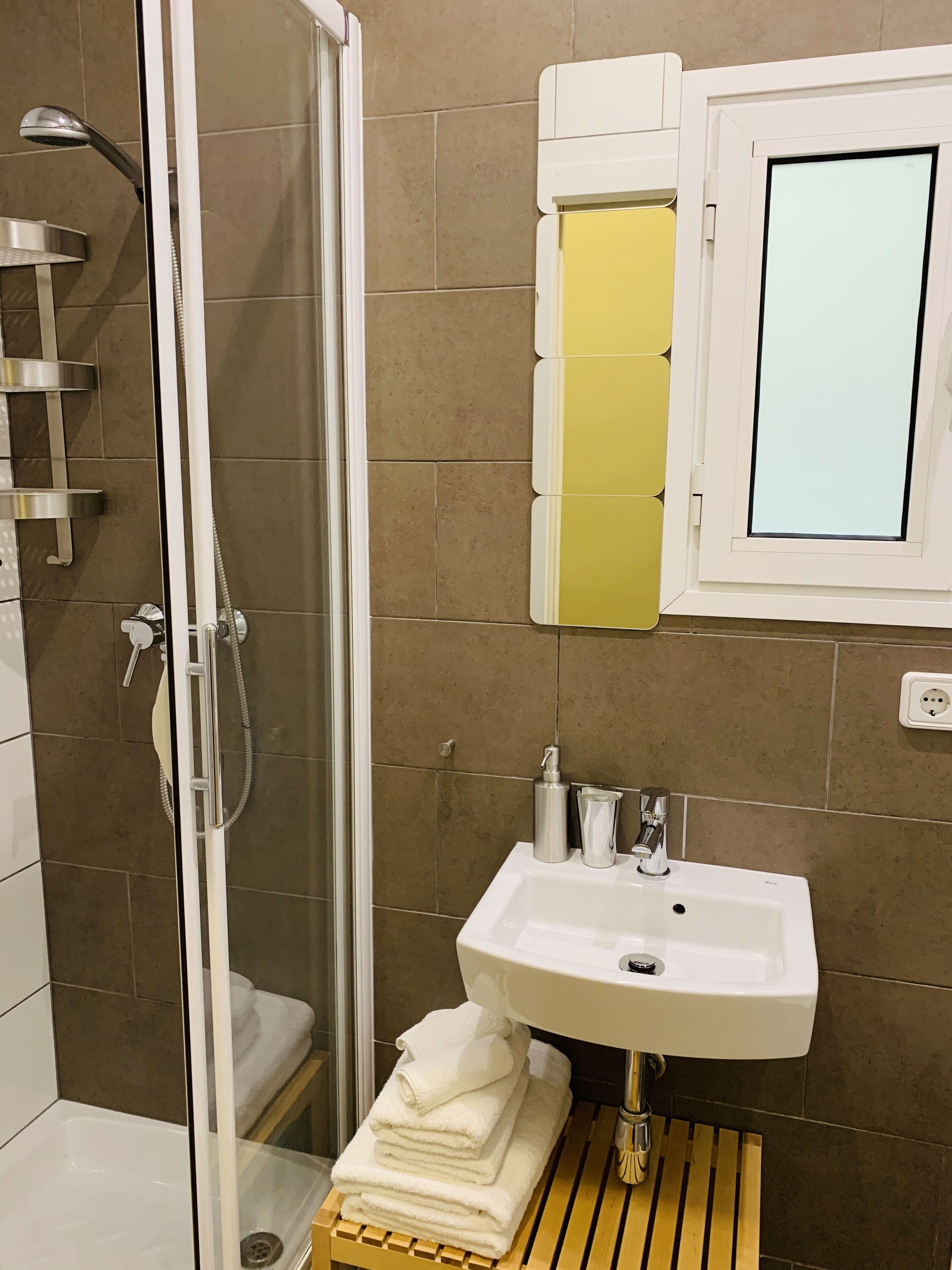 Un baño con ducha de vidrio, un lavamanos con espejo y una pila de toallas en un estante de madera.