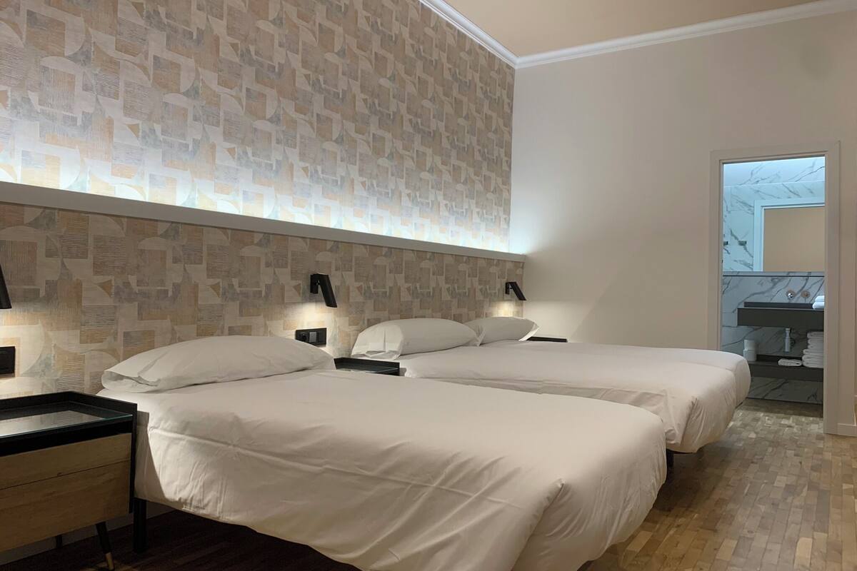A hotel room with two beds, a nightstand, a wall-mounted lamp, and a bathroom visible through an open door.