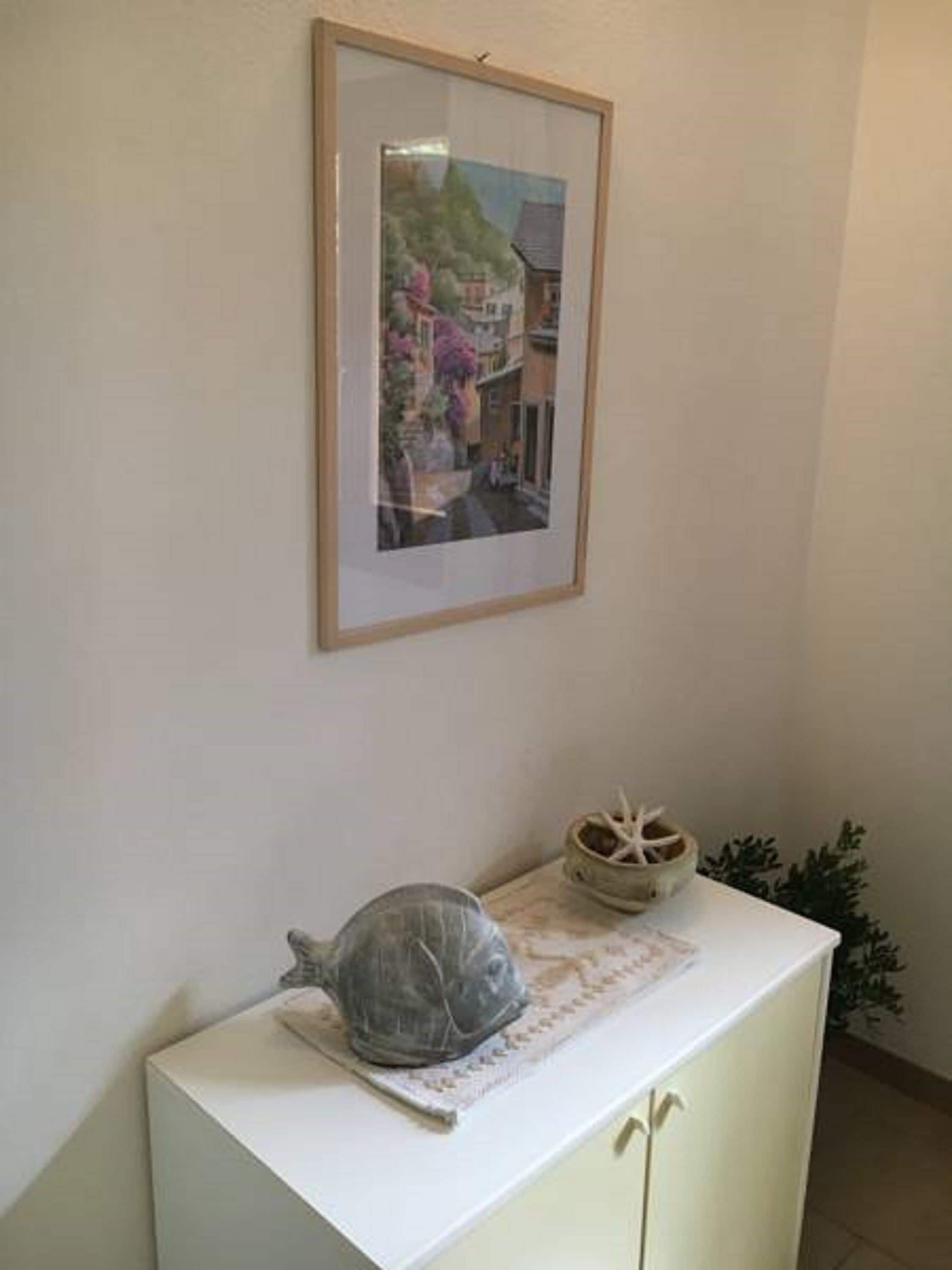 Un tableau encadré représentant une scène de rue, accroché au mur, une petite table sur laquelle se trouvent un bol décoratif et une figurine, et un meuble avec un tiroir.