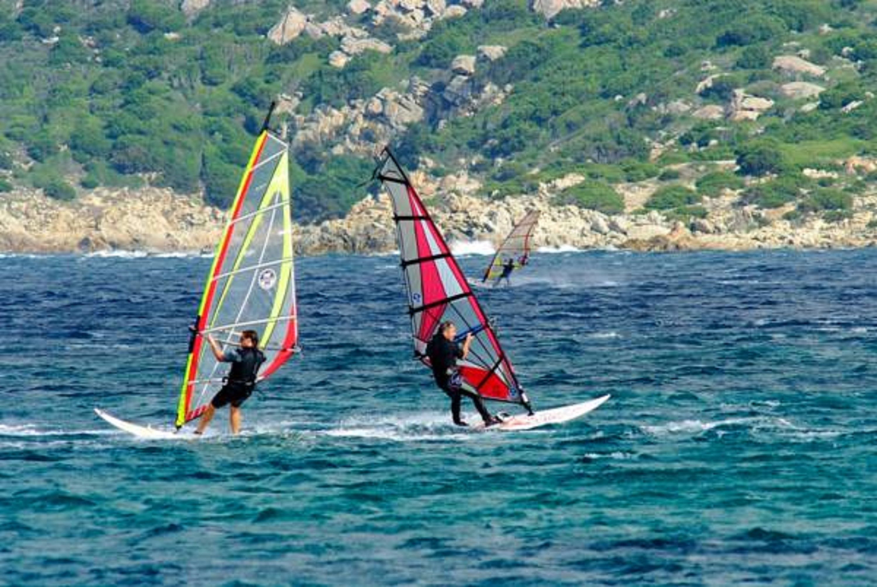 Zwei Windsurfer auf dem Wasser, im Hintergrund eine felsige Küste und viel Grün.