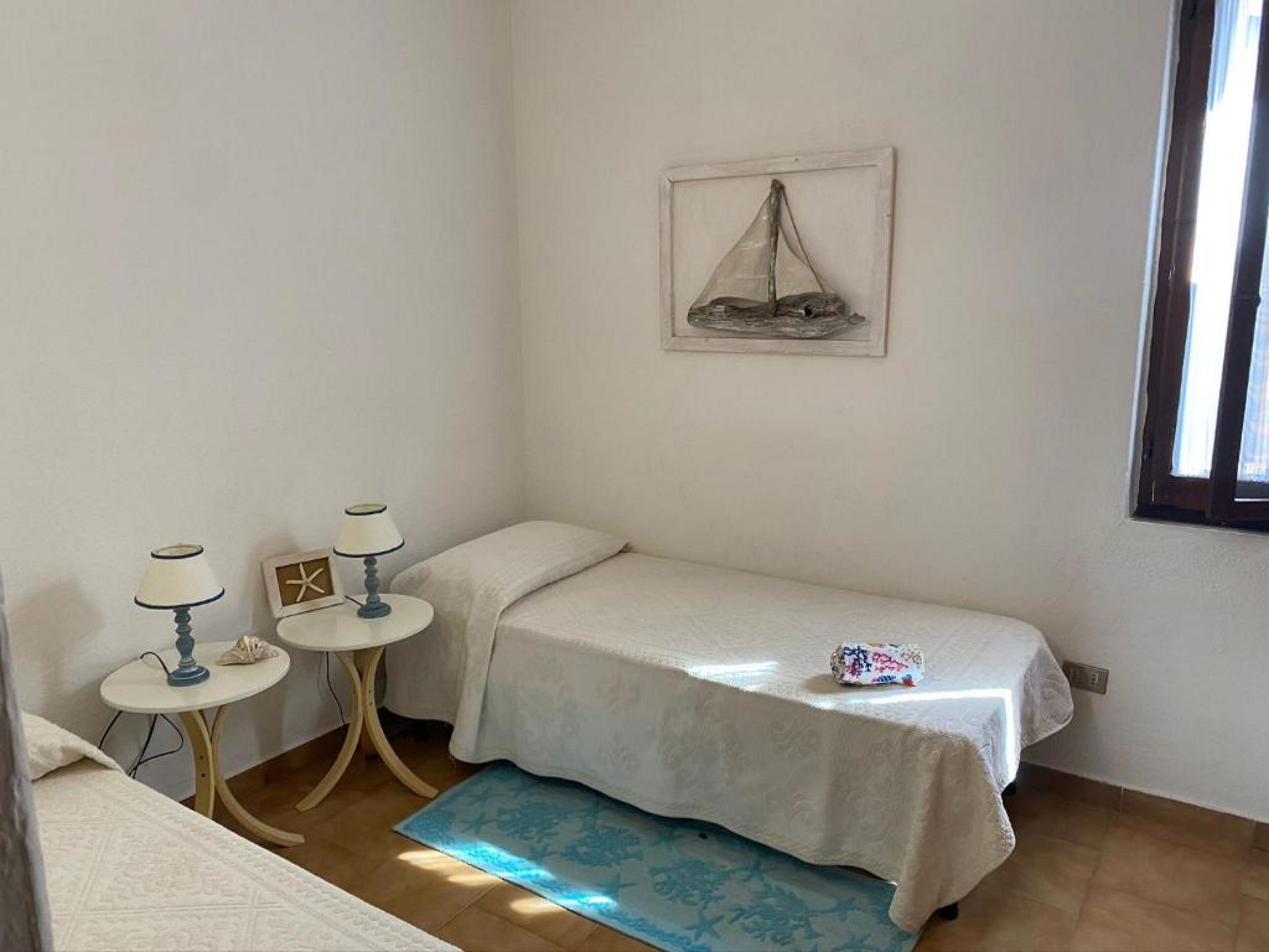 A small, clean room with a single bed, two bedside tables, a small table with a lamp, a framed picture on the wall, and a window.