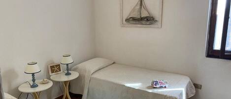A small, clean room with a single bed, two bedside tables, a small table with a lamp, a framed picture on the wall, and a window.