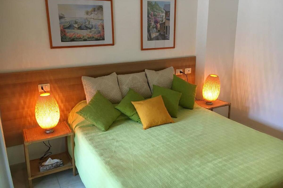 A neatly made bed with green and yellow pillows, two bedside lamps, and two framed pictures on the wall.