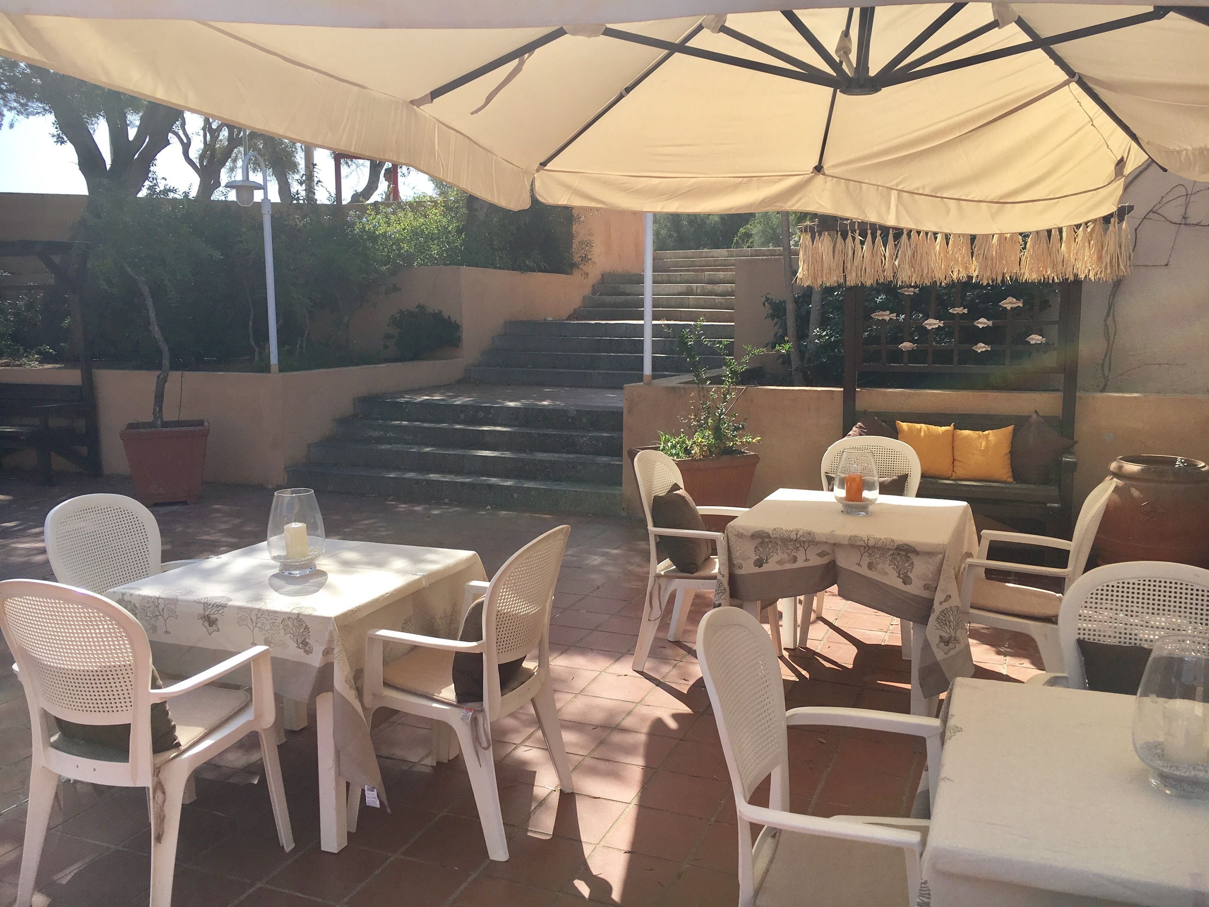An outdoor patio with white plastic chairs and tables, a beige umbrella, and a staircase leading to a higher level.