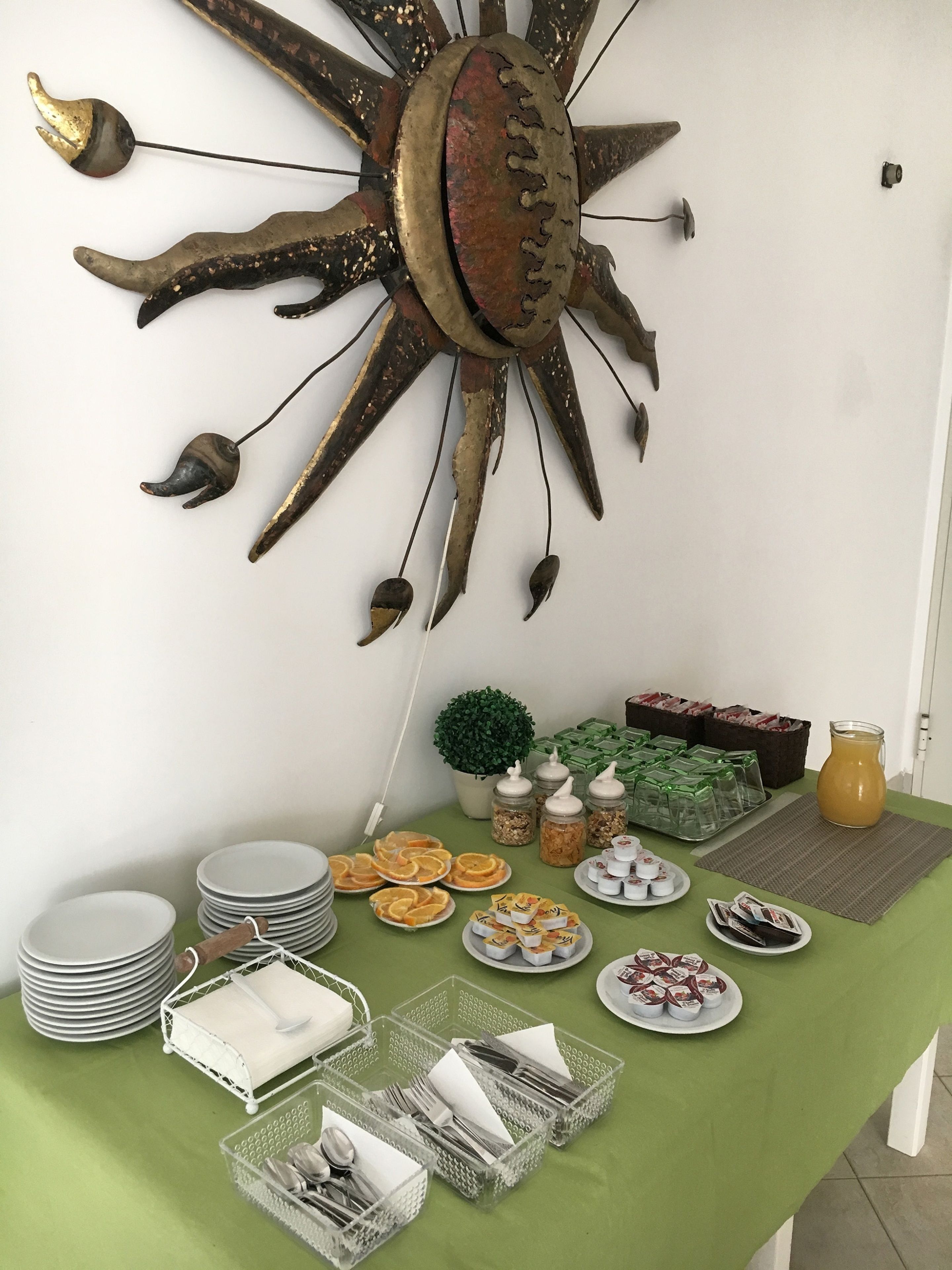 Una mesa dispuesta con platos, cubiertos y alimentos, un adorno de pared con forma de sol, y una pequeña planta en maceta.
