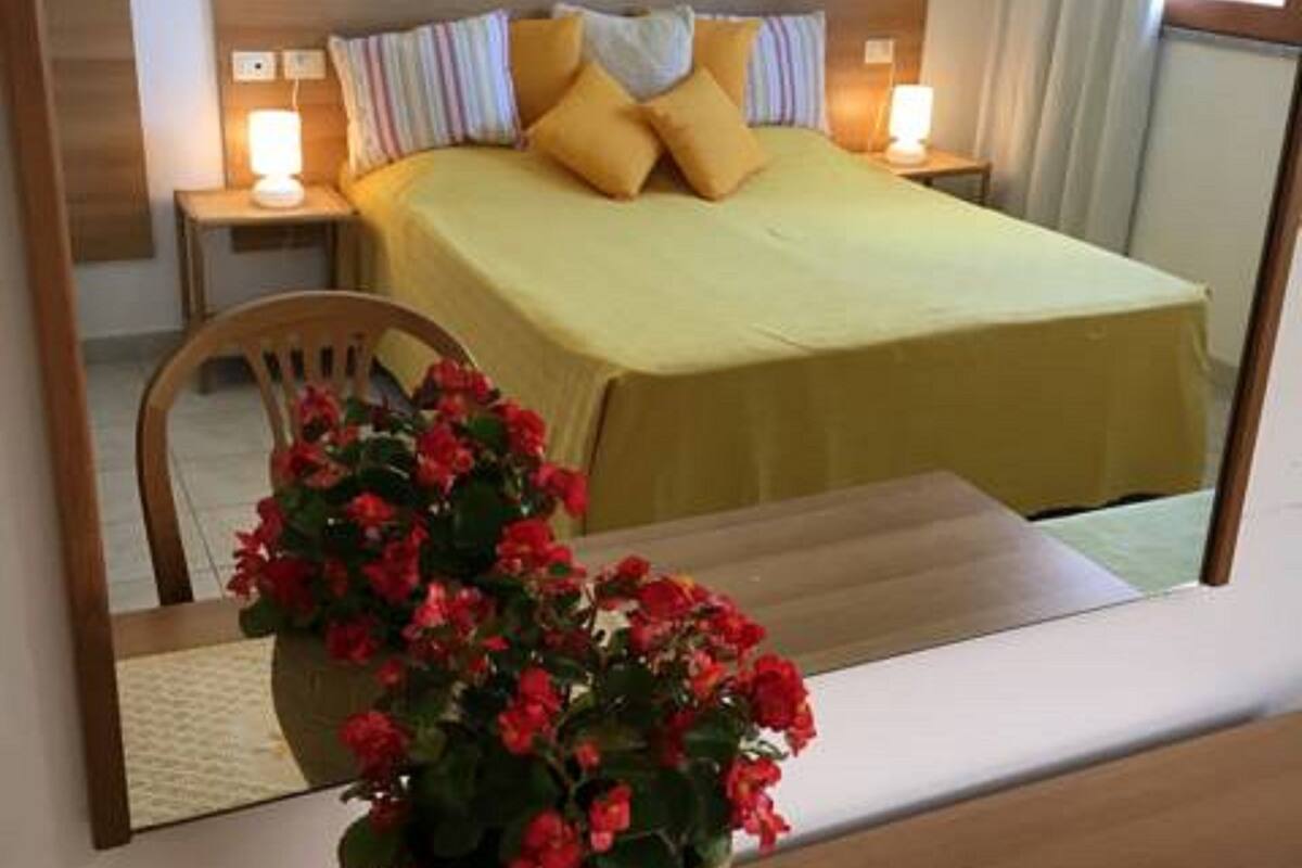 A hotel room with a bed, bedside lamps, a painting on the wall, and a potted plant with red flowers.