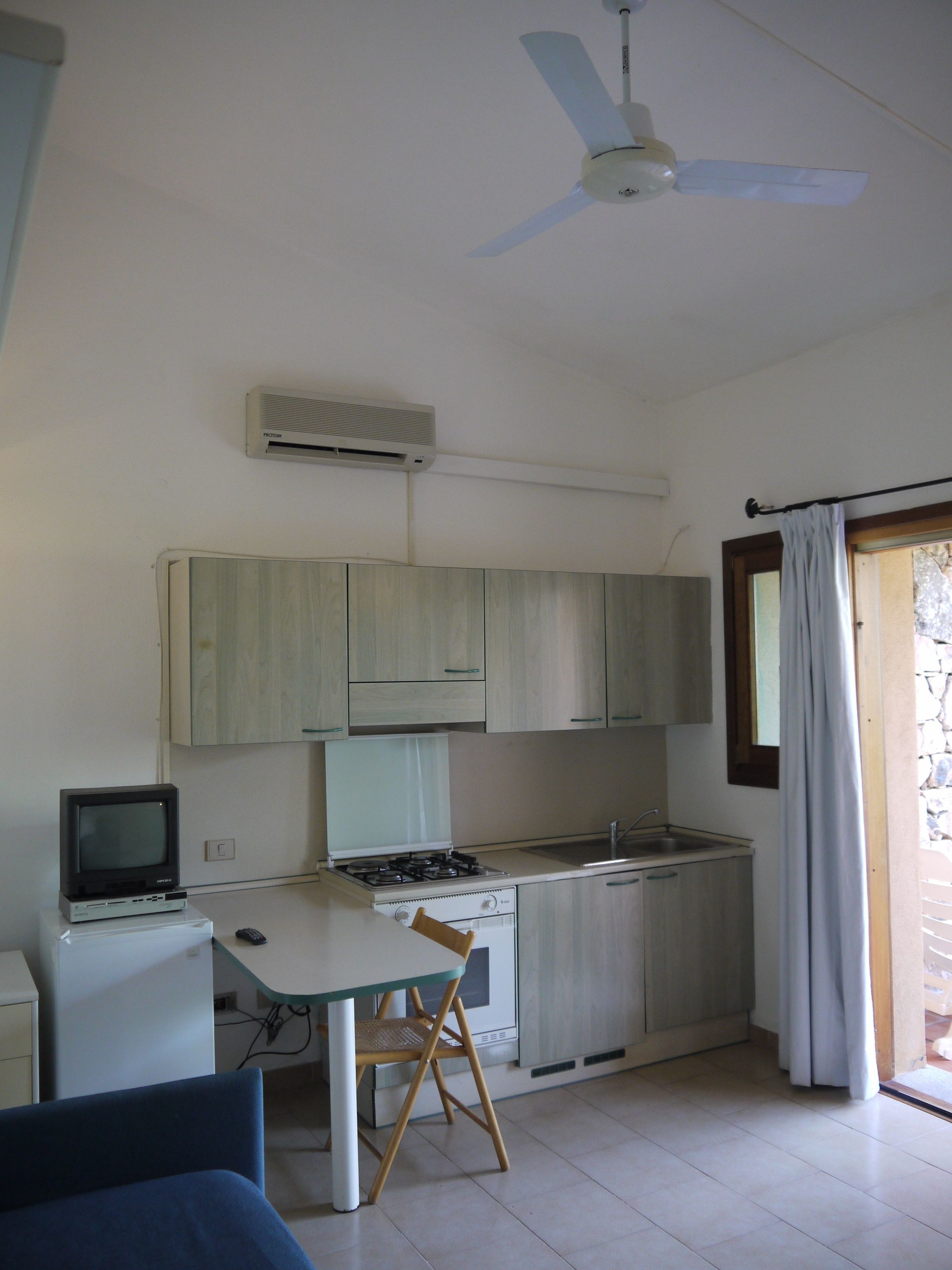 A compact kitchen with a gas stove, a small refrigerator, a television, and a ceiling fan.