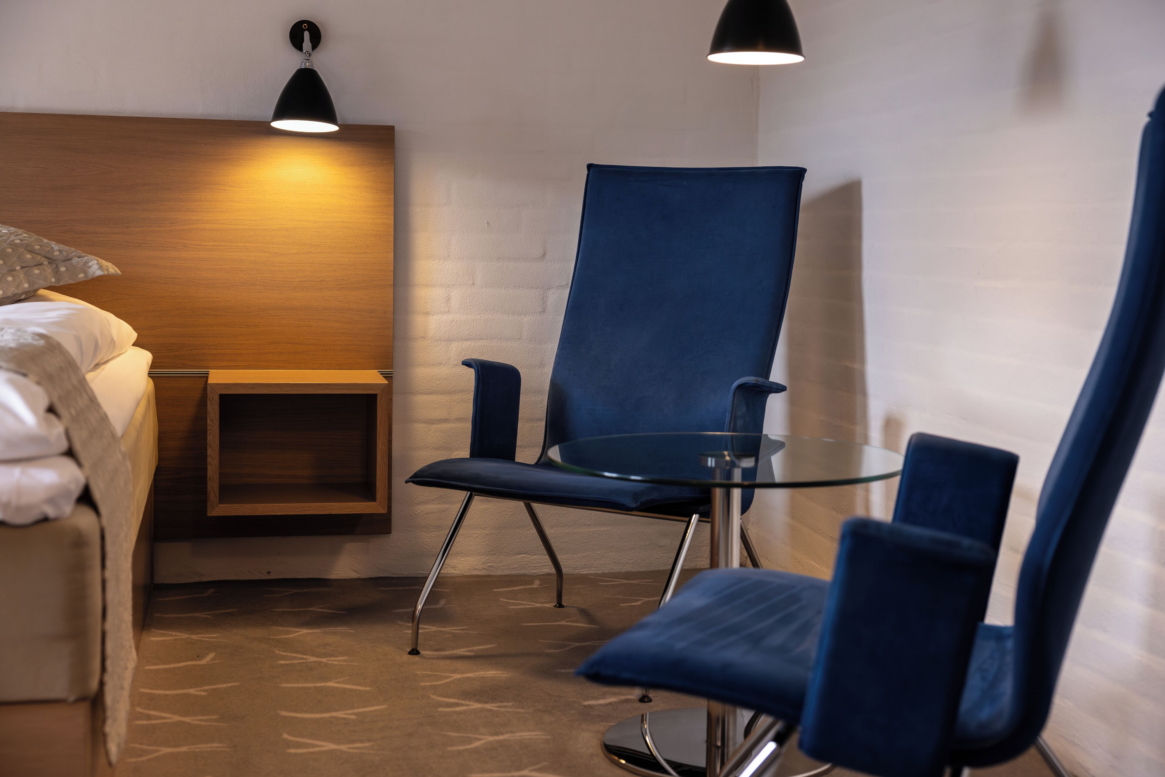 Hotelkamer met een bed, een houten hoofdbord, een blauwe stoel, een glazen tafel en een wandlamp.
