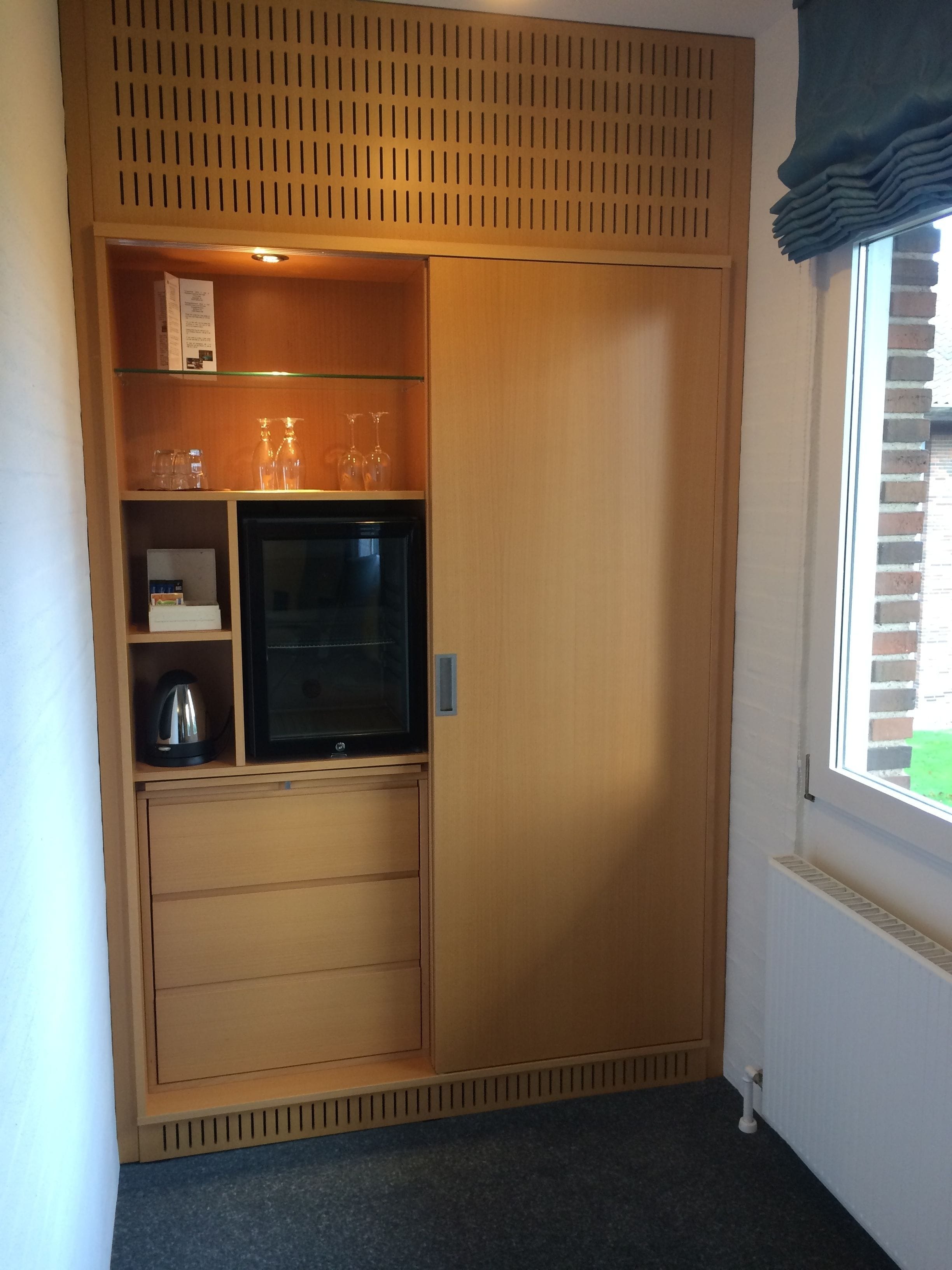 Una habitación compacta con televisión integrada, tetera y un pequeño estante con vasos.
