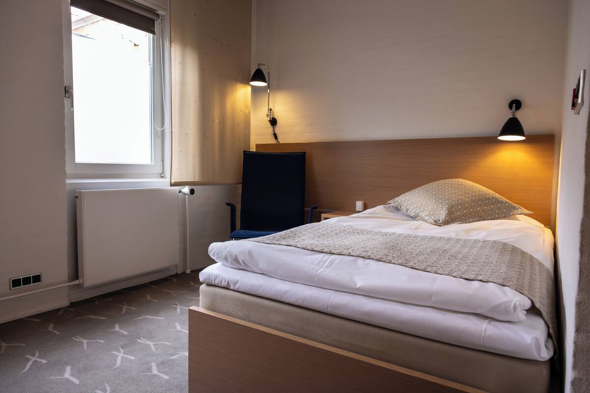 Una cama bien hecha con sábanas blancas, cabecera de madera y dos lámparas de lectura fijadas a la pared.