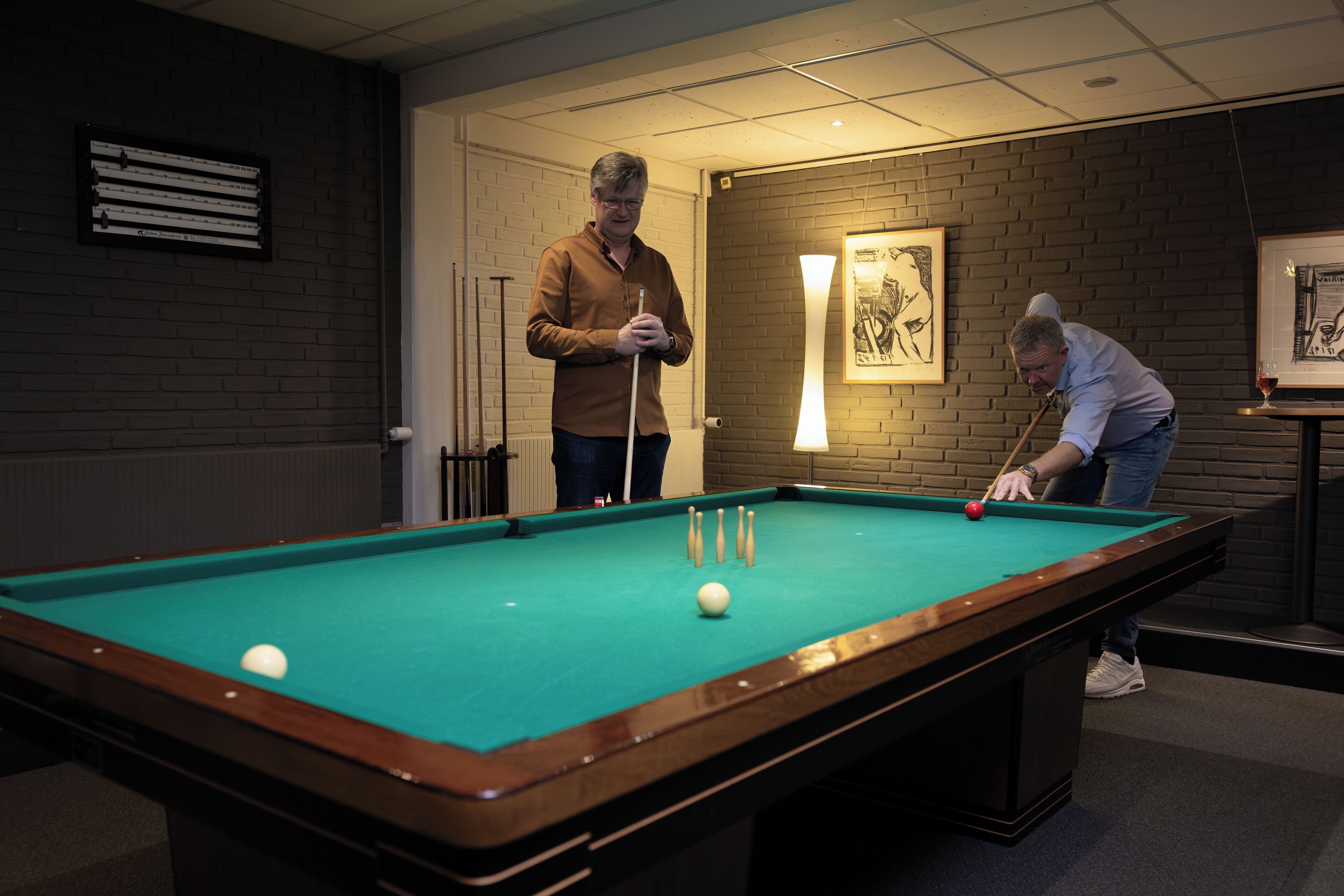 A pool table with two players, a framed picture on the wall, and a lamp.