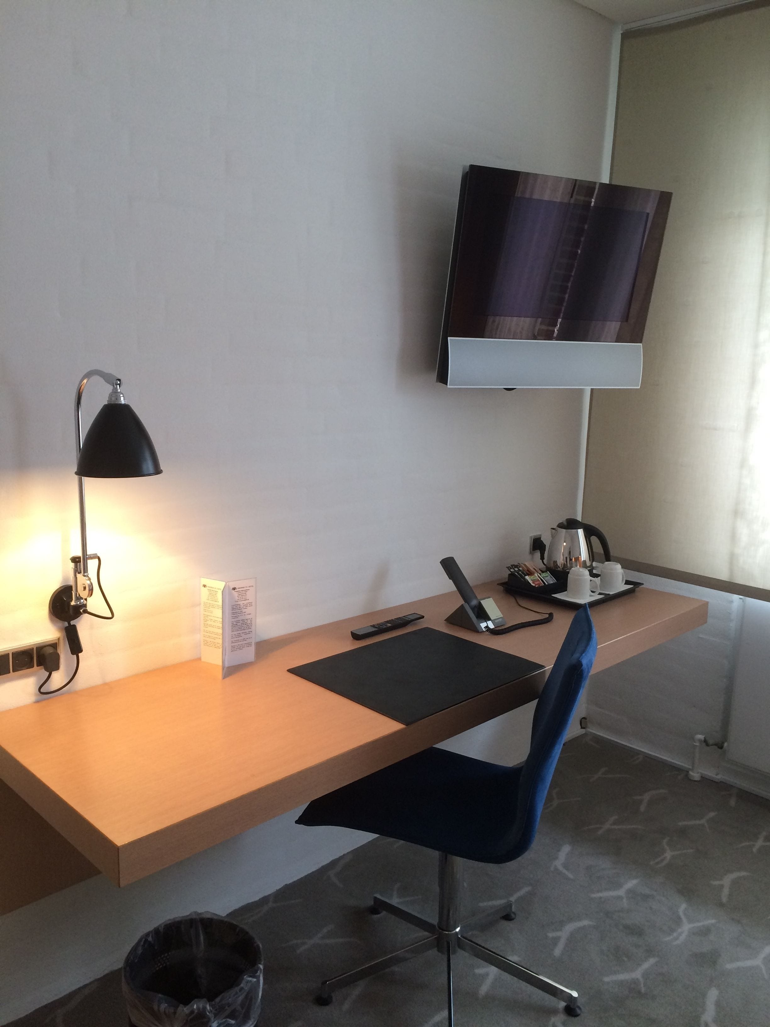Habitación de hotel con un escritorio de madera, una silla de oficina negra, una lámpara de escritorio, un televisor de pantalla plana montado en la pared, una cafetera y un cubo de basura.