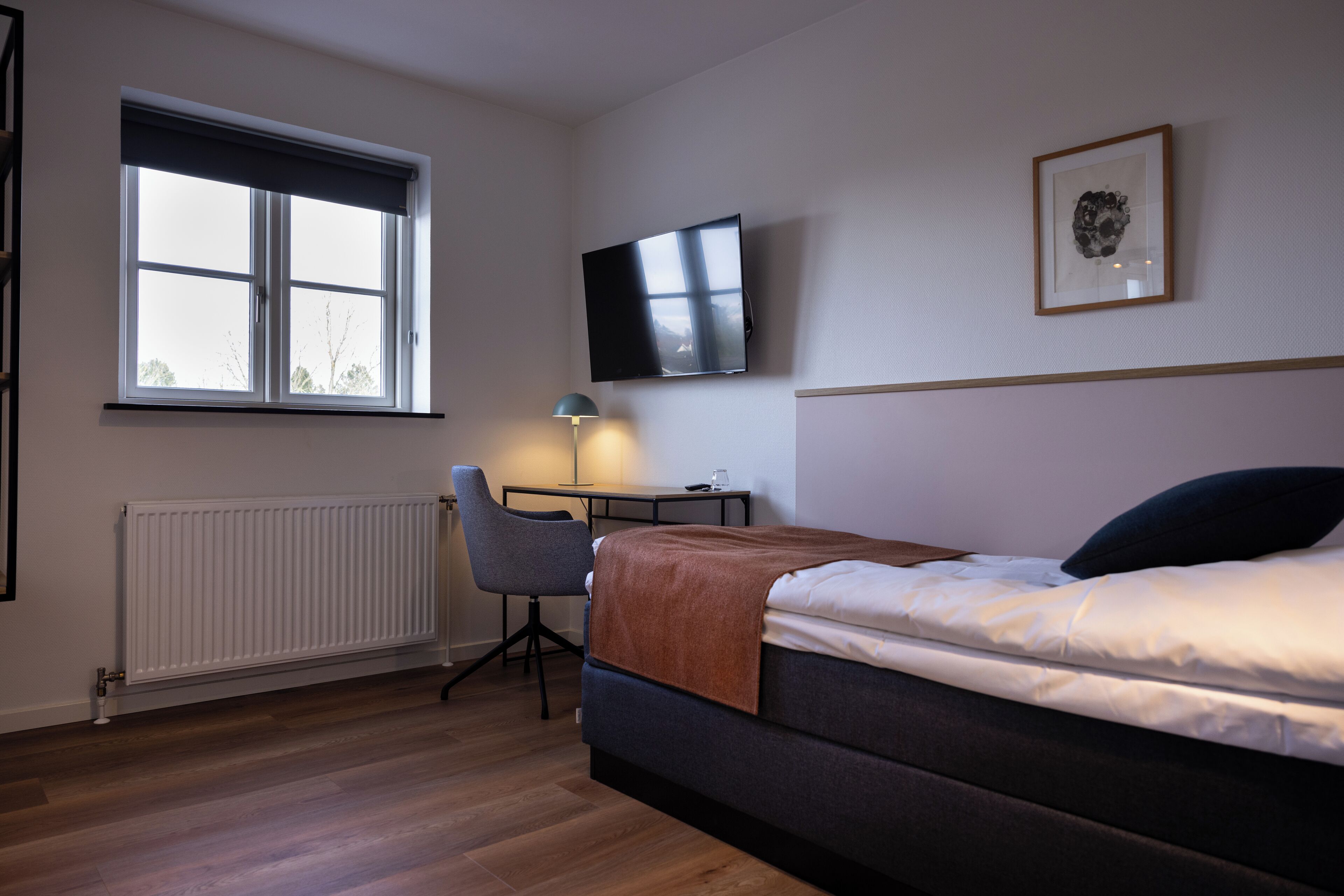 A bedroom with a bed, a desk, a chair, a radiator, a TV, and a window.