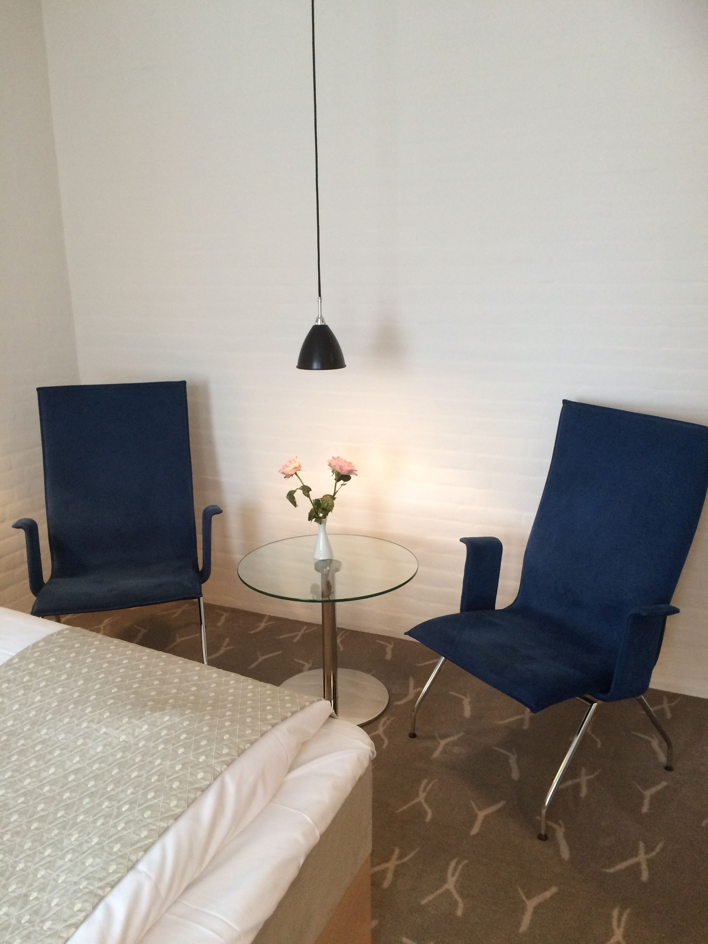 Habitación de hotel con una cama, dos sillas azules, una mesita de vidrio con un jarrón de flores y una lámpara colgante.