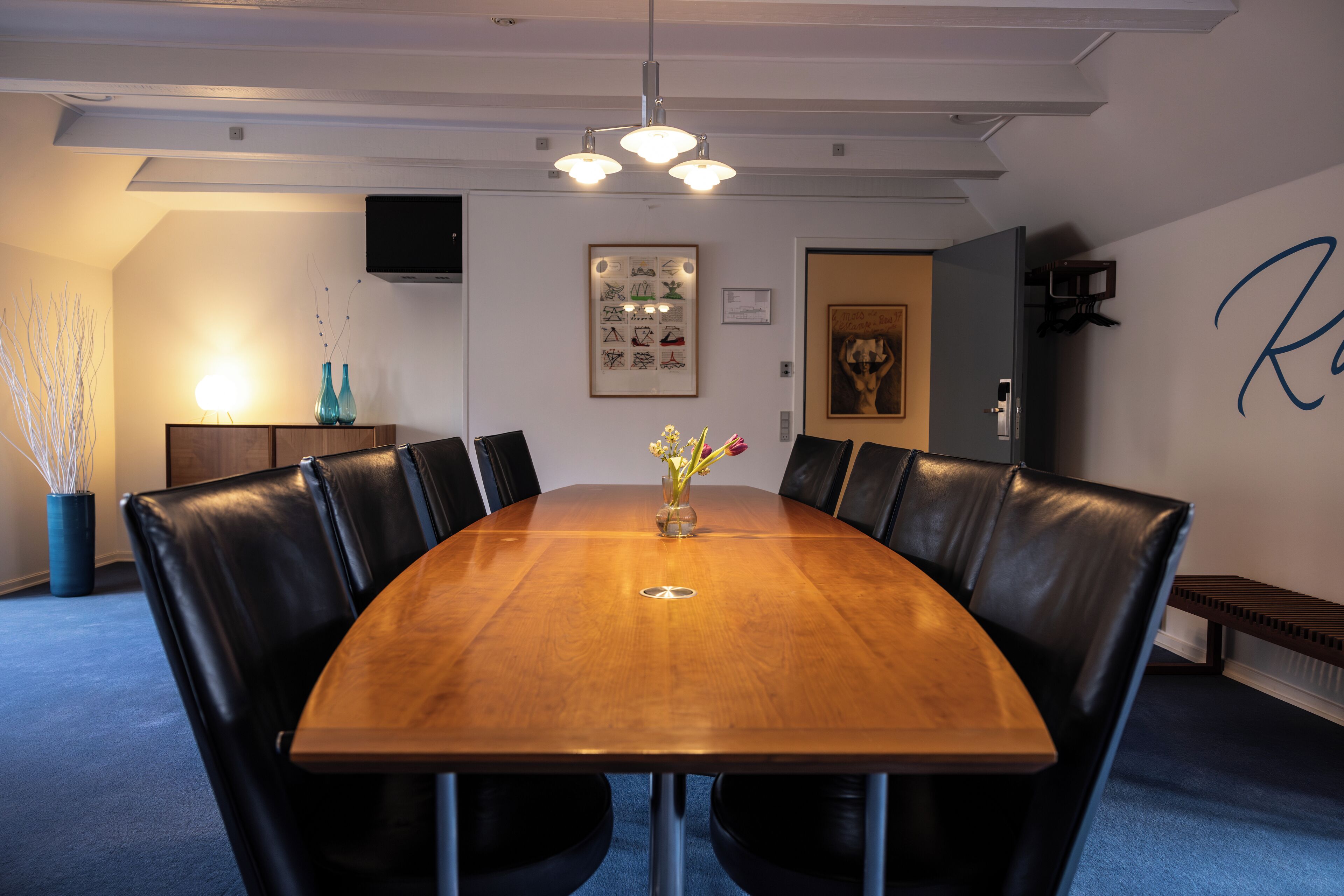 Un salón de conferencias con una mesa de madera, sillas de cuero negro, un jarrón con flores, un cuadro enmarcado y una pared con una letra grande.