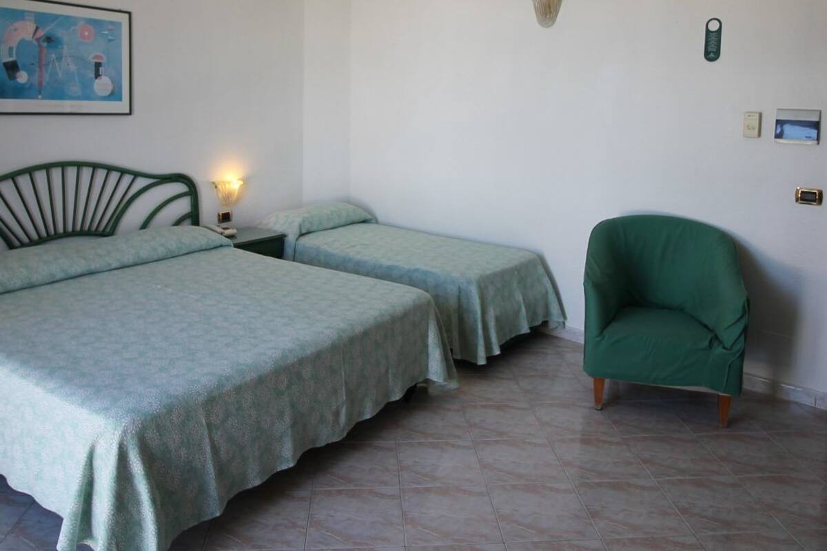 Una habitación con dos camas individuales, un sillón verde, una mesita de noche con lámpara y un reloj de pared.