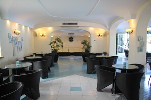 A hotel lobby with round tables and wicker chairs, a clock on the wall, and a tiled floor. - Hotel Park Imperial (Forio)