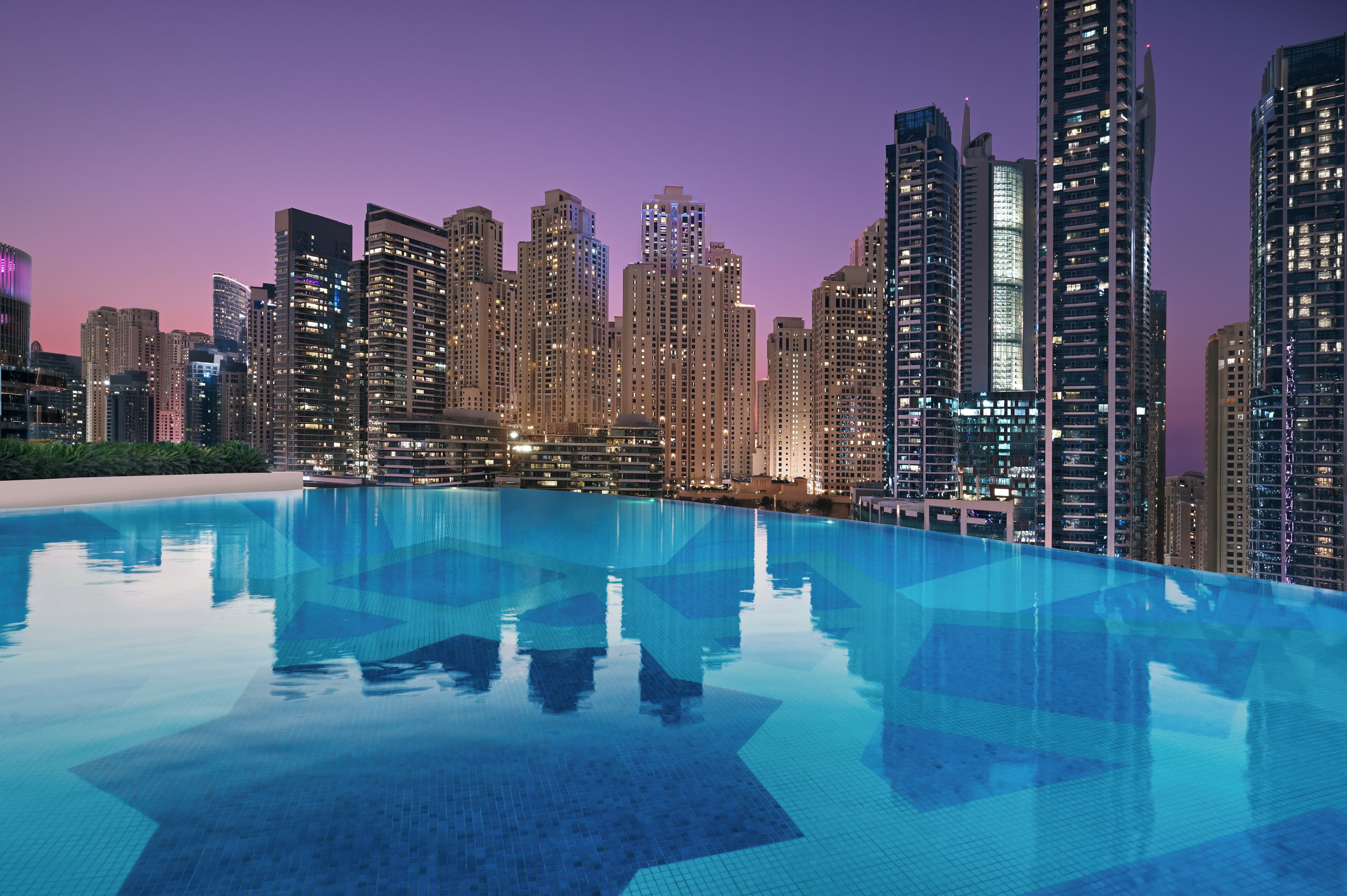A modern hotel with a rooftop infinity pool overlooking a city skyline at dusk.