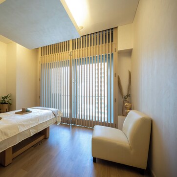 A minimalist room with a bed, a chair, a small table, and a window with blinds.