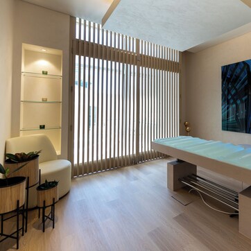 A modern room with a glass-top table, a sofa, a chair, a wall-mounted shelf, and a large abstract painting.
