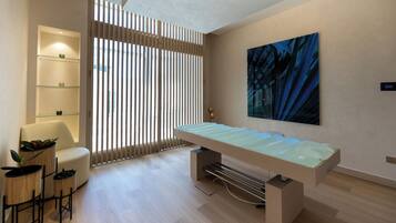A modern room with a glass-top table, a sofa, a chair, a wall-mounted shelf, and a large abstract painting.