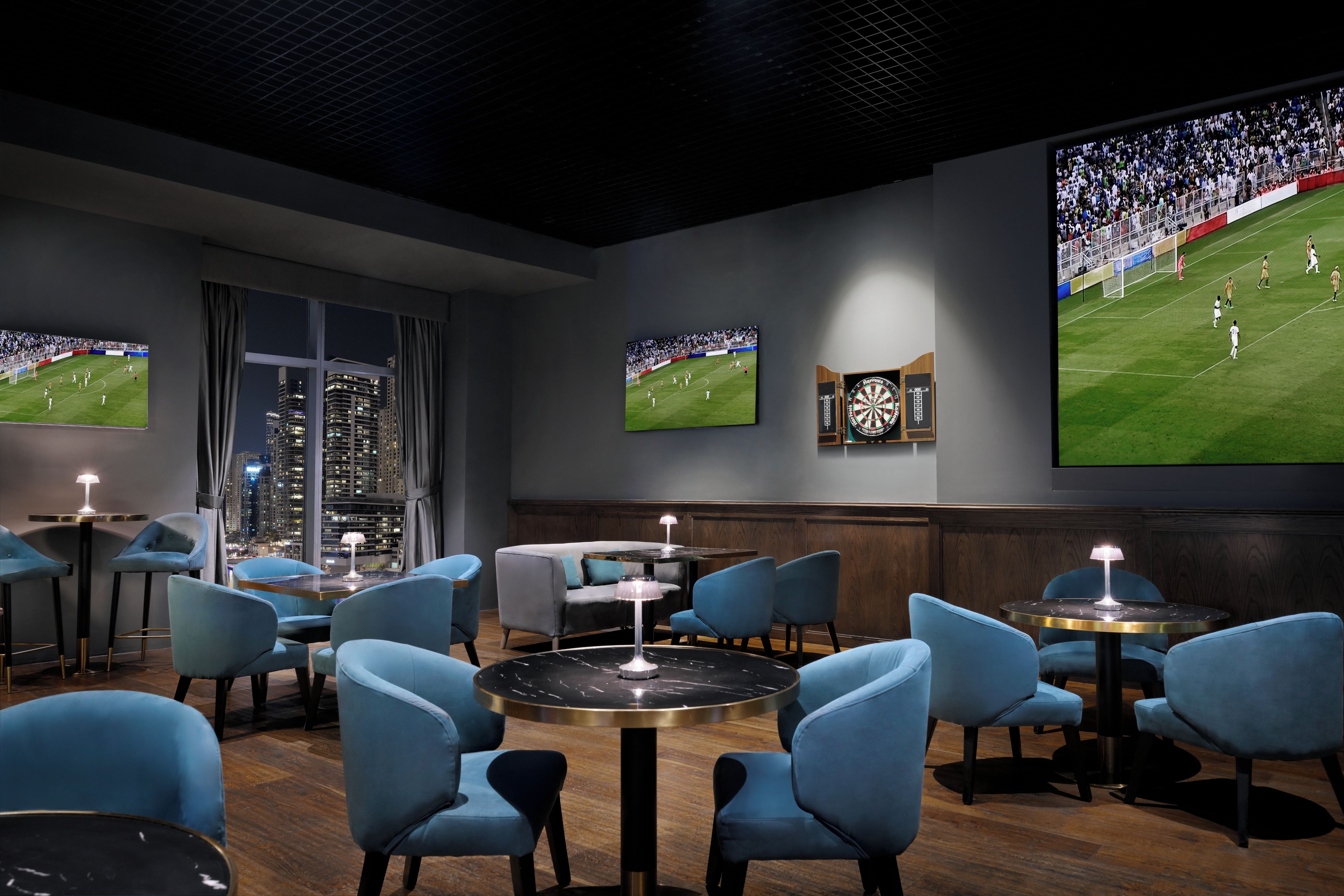 A modern lounge with multiple seating areas, a dartboard, and large screens displaying sports events.