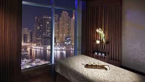 Une chambre d’hôtel dotée d’une grande fenêtre donnant sur une vue panoramique de la ville la nuit, d’un lit avec un plateau d’articles de toilette et d’une petite étagère ornée d’objets décoratifs.