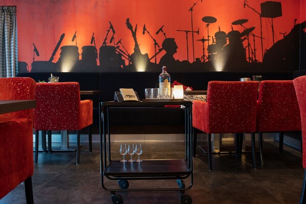 Un área de bar con sillas rojas, un carrito con vasos y un mural en la pared de músicos.