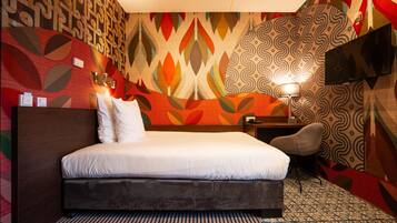 A hotel room with a large bed, a desk, a chair, a TV, and a patterned wall.