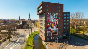 A modern building with a large mural on its side, a parking area, and a canal in the foreground. - Hampshire Delft Centre (Delft)