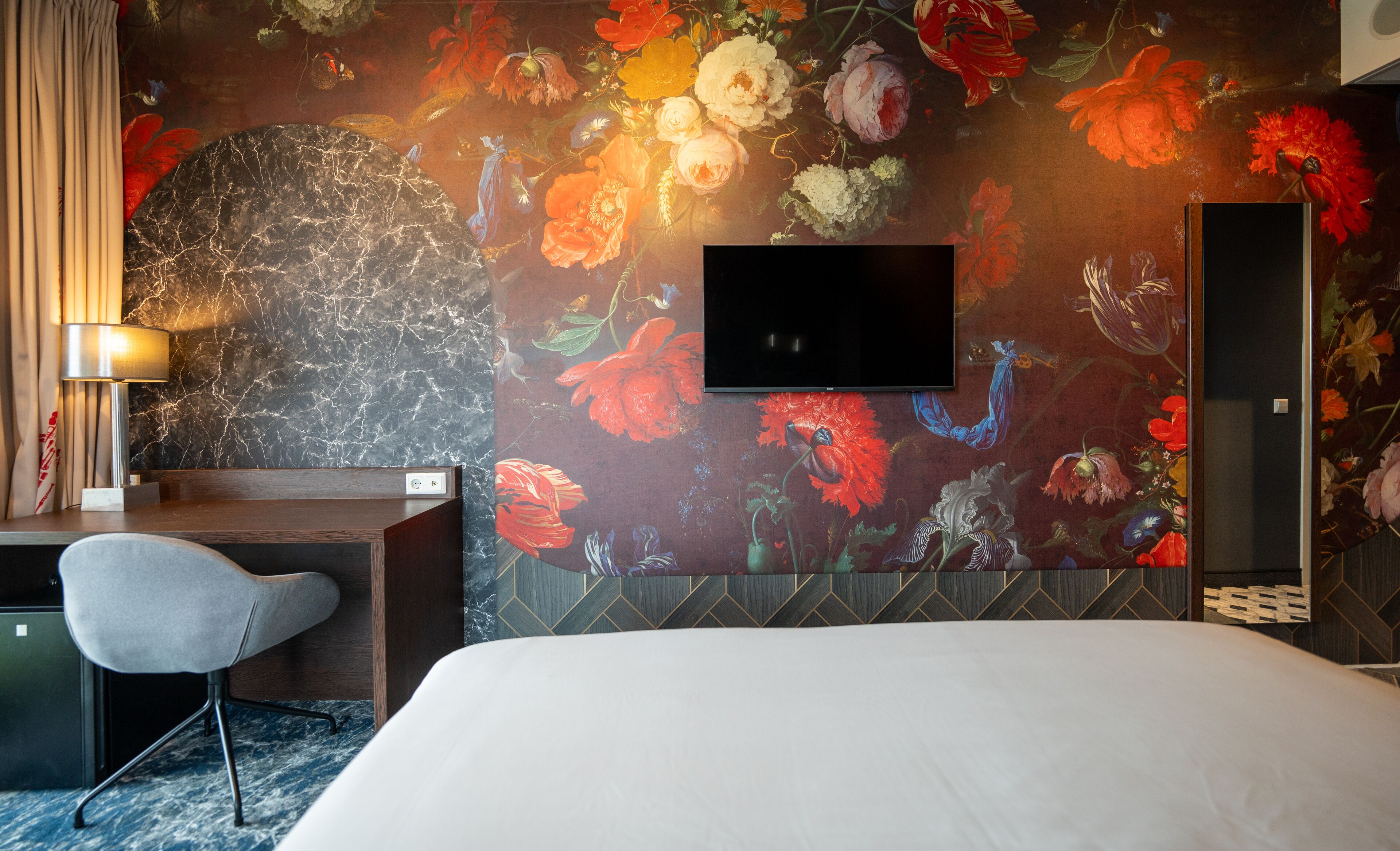 Un dormitorio de hotel con una cama grande, un escritorio con silla, un televisor de pantalla plana montado en la pared y un mural floral.