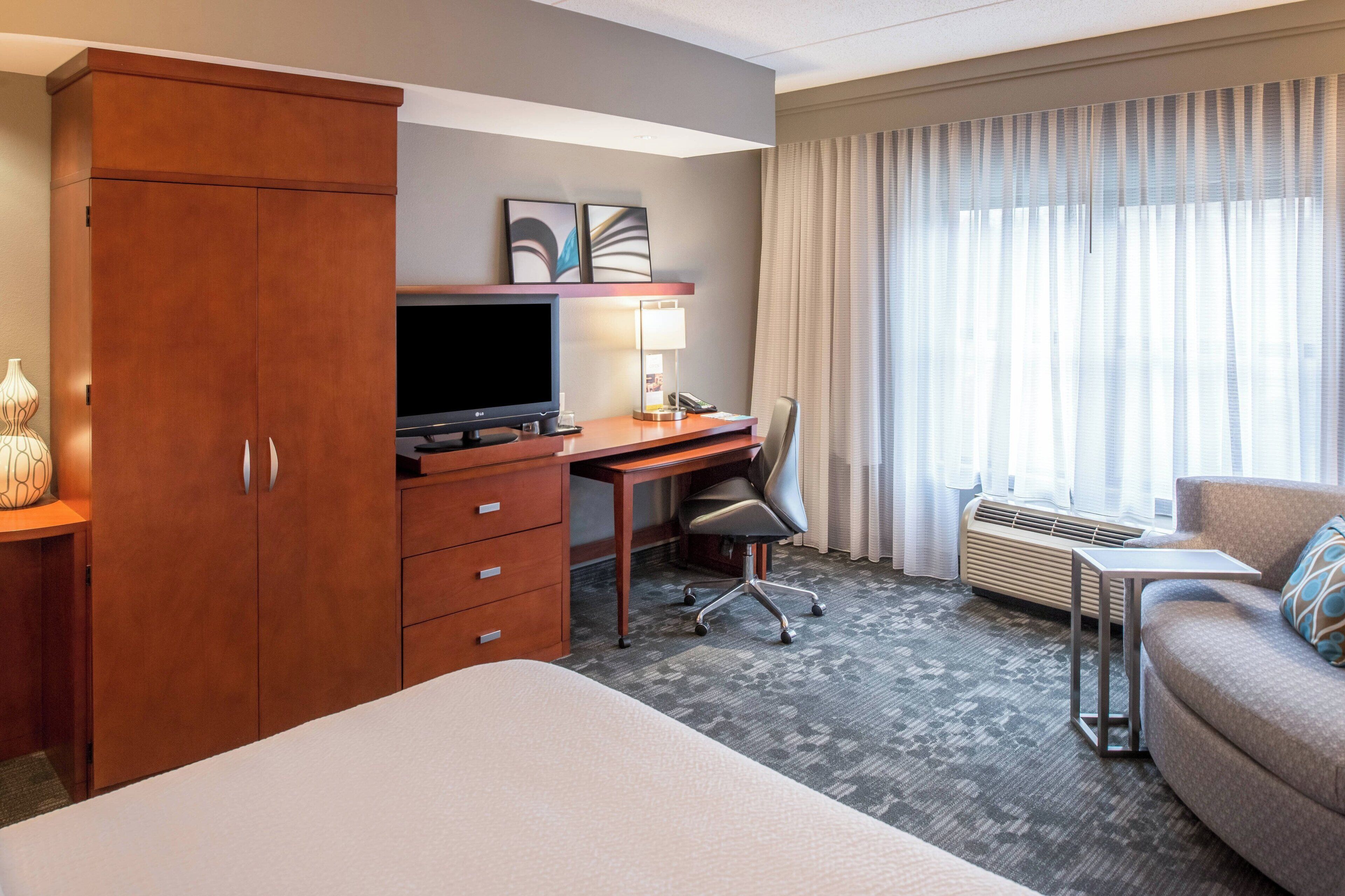 Ein Hotelzimmer mit Bett, Schreibtisch mit Computer, Stuhl, Sofa, Kleiderschrank und einem Fenster mit Vorhängen.
